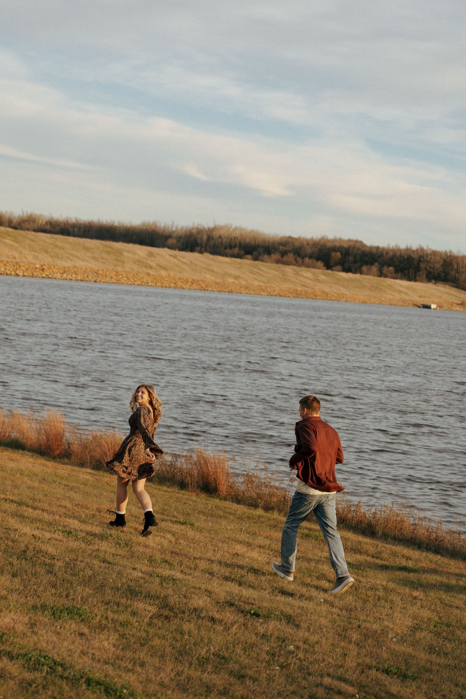 People walking along the grassy shoreline of a lake during sunset.