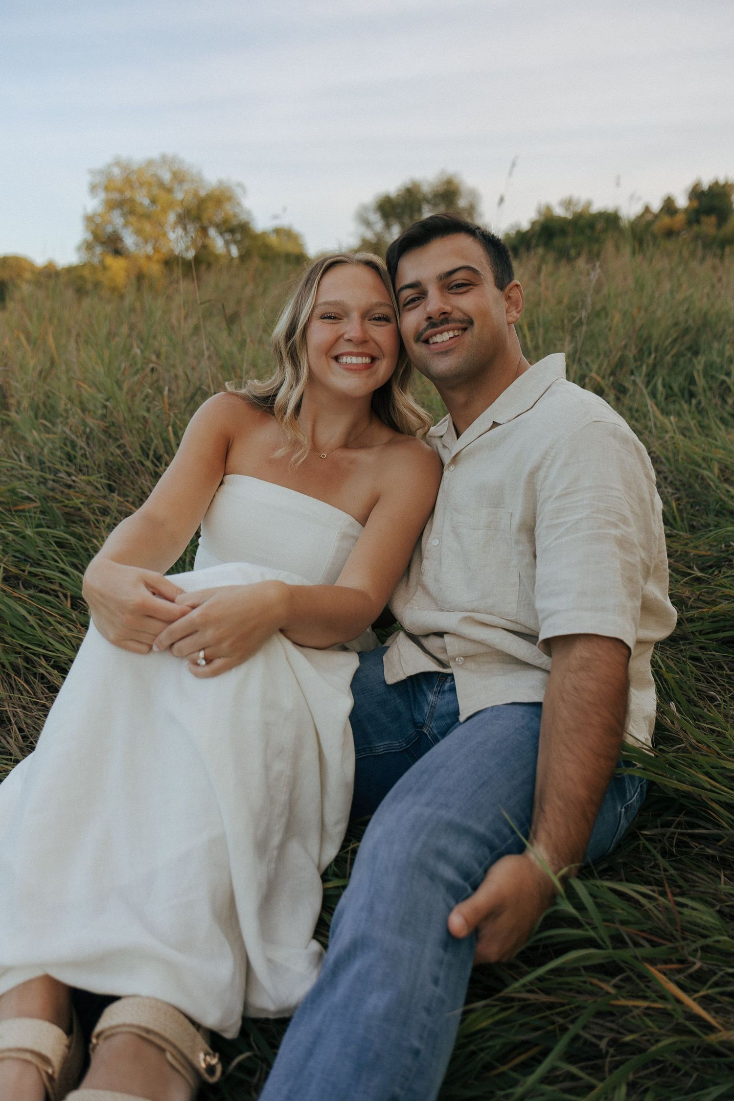 A joyful couple laughs together while sitting in tall grass during their outdoor photoshoot.