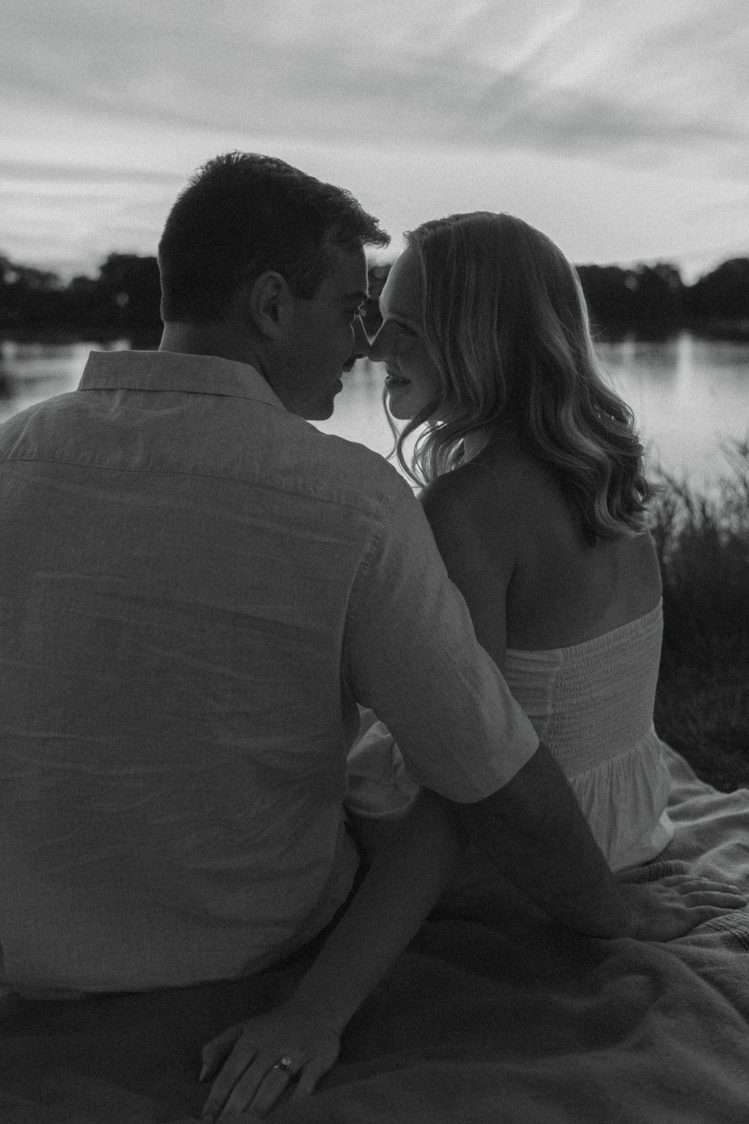 A romantic black and white photo series of a couple sharing intimate moments by a lake at sunset.