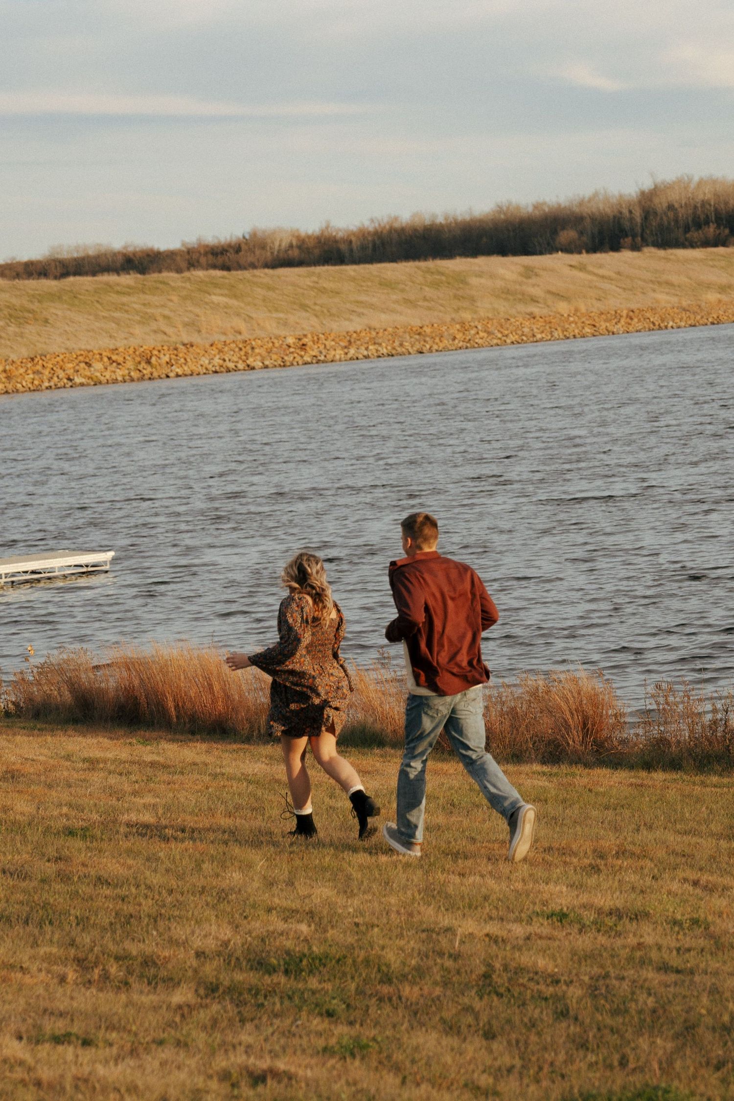 Couple running playfully along the lakeside grass during a golden autumn evening.