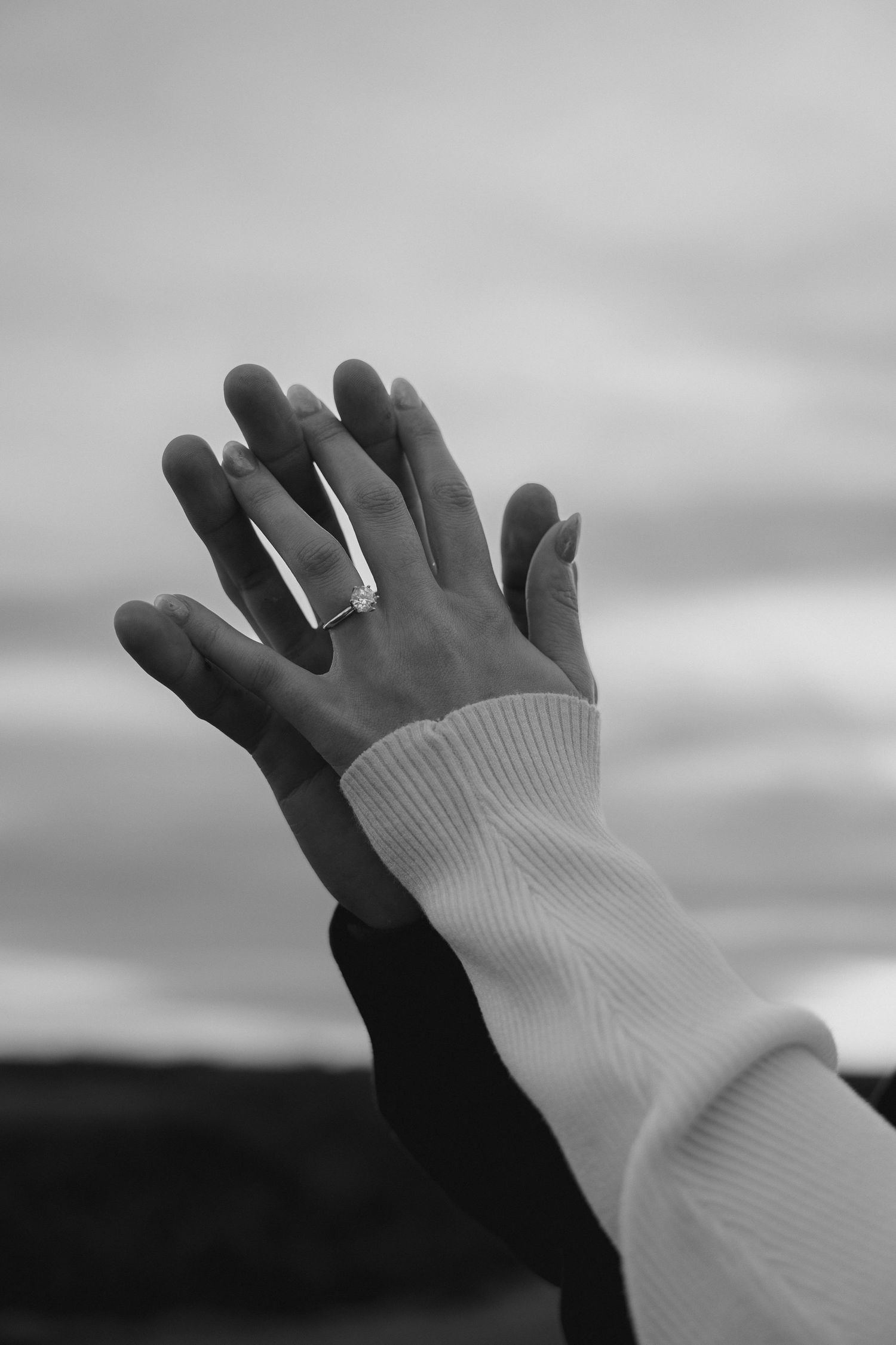 A black and white image of clasped hands with an engagement ring against the sky.
