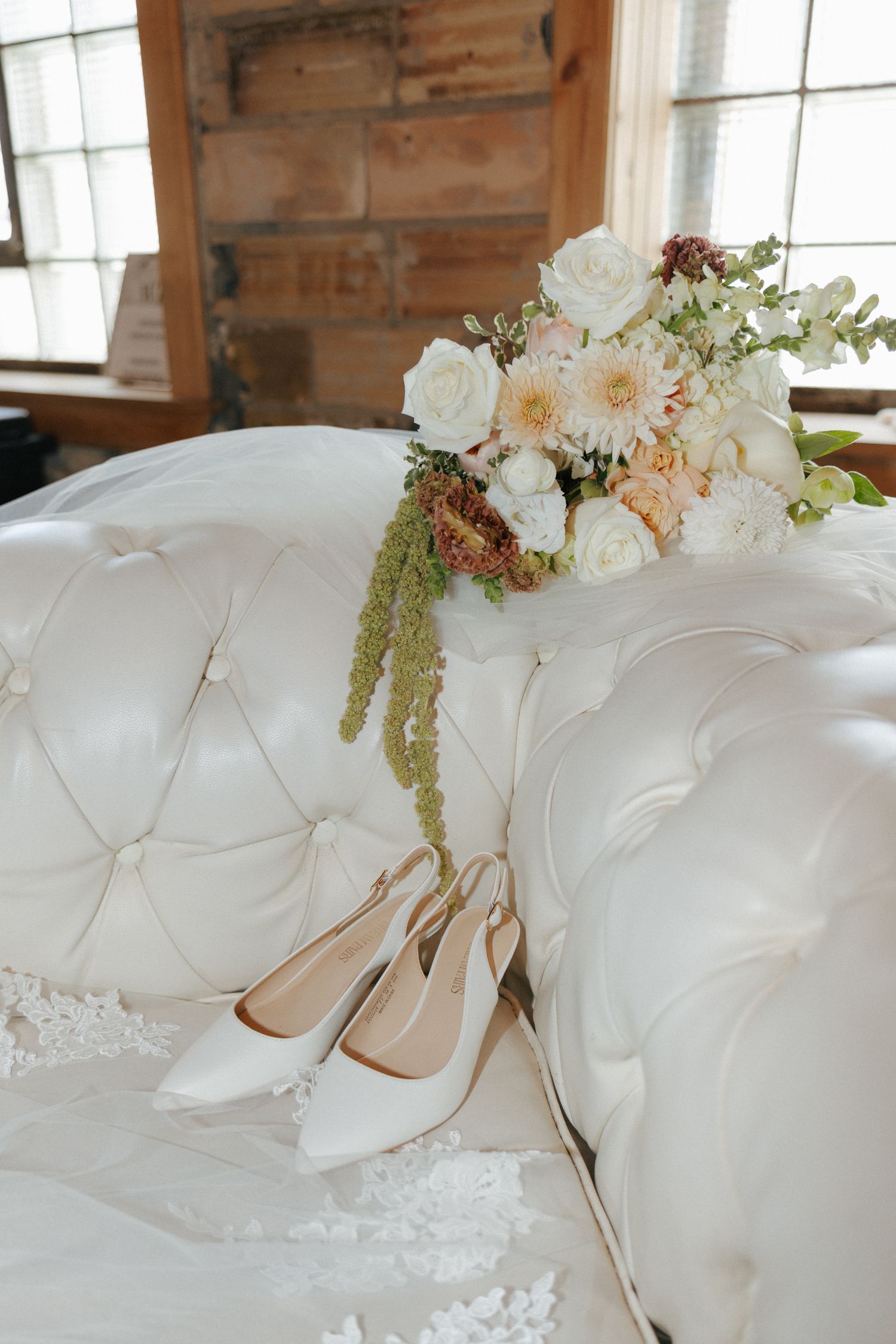 Elegant bridal heels and a white floral bouquet with peach accents rest on a tufted white velvet sofa.