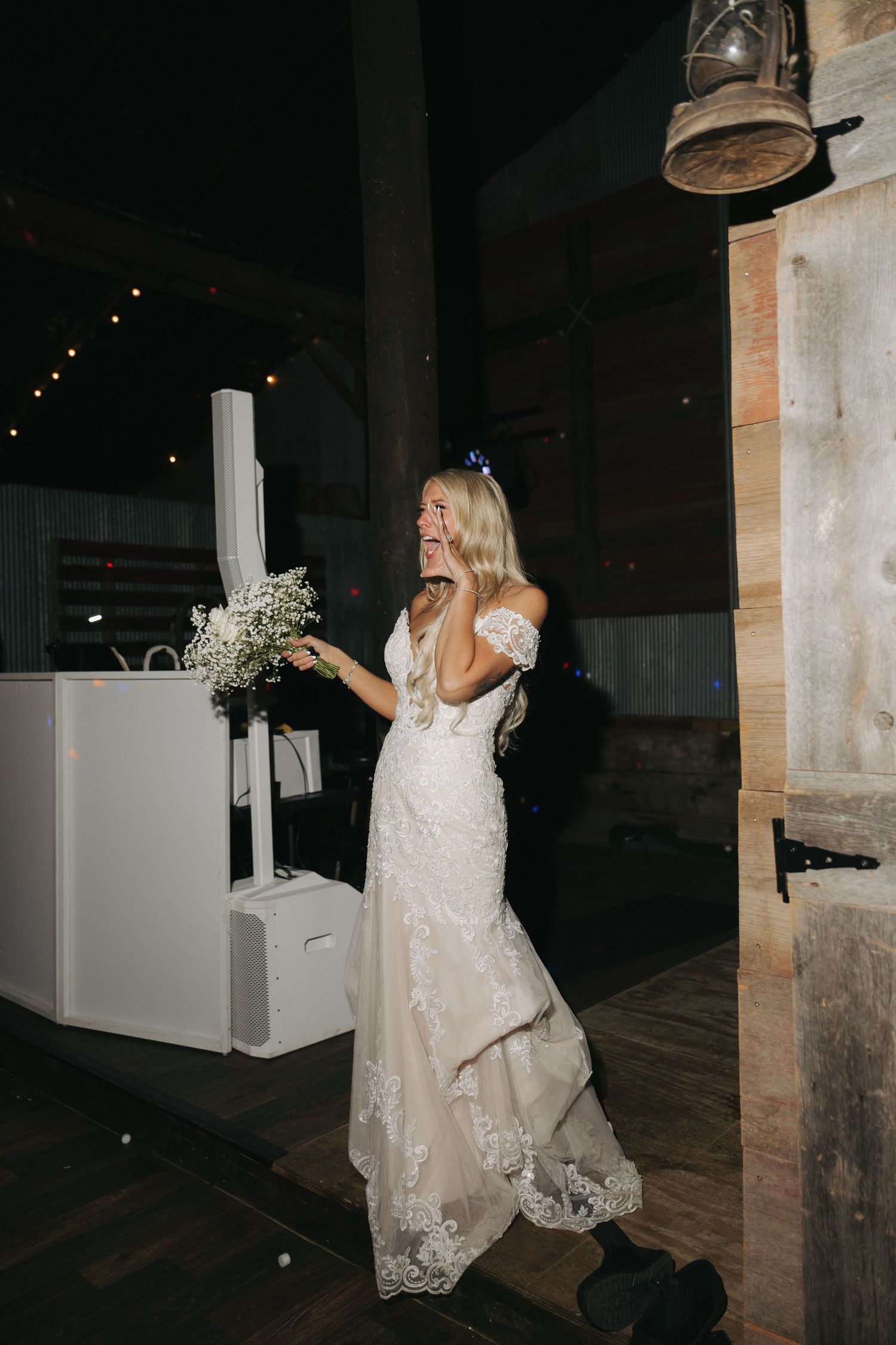 Radiant bride in lace off-shoulder gown laughs while holding a white bouquet inside a rustic barn venue.