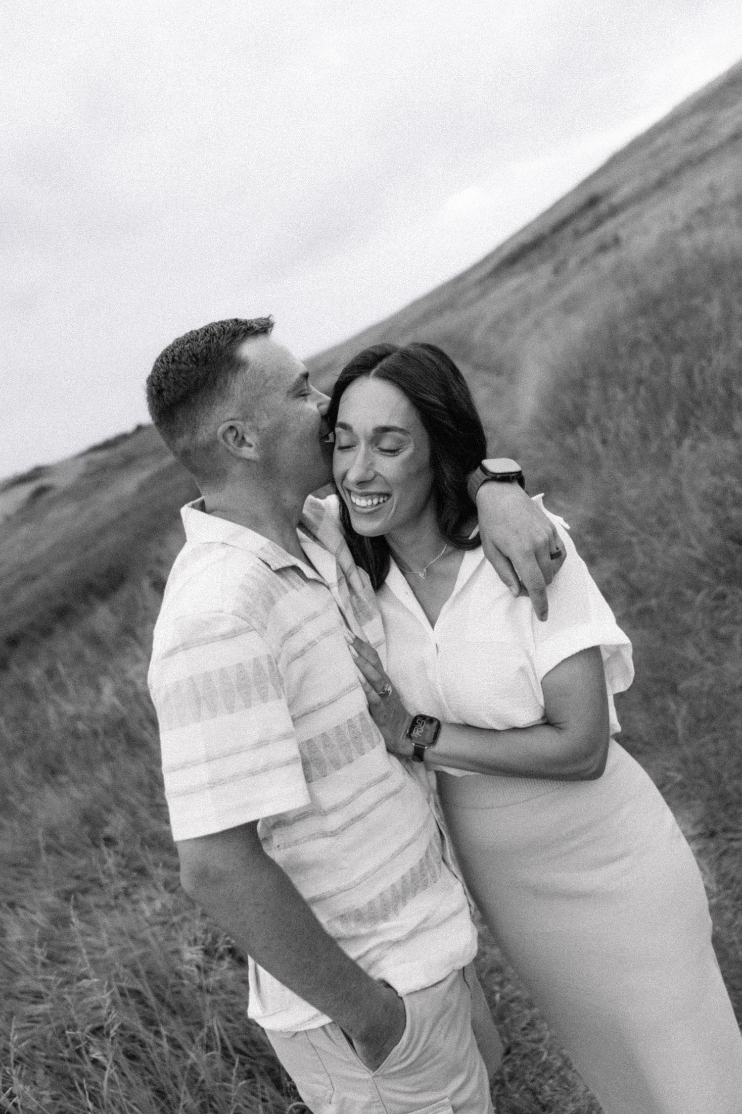 Black and white photo of a couple embracing on a hillside, man whispering to smiling woman outdoors.