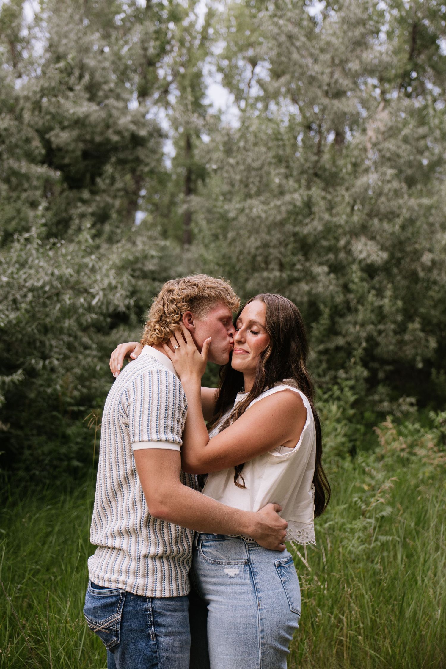 A couple shares a romantic kiss surrounded by lush green trees and tall grass in an outdoor setting.