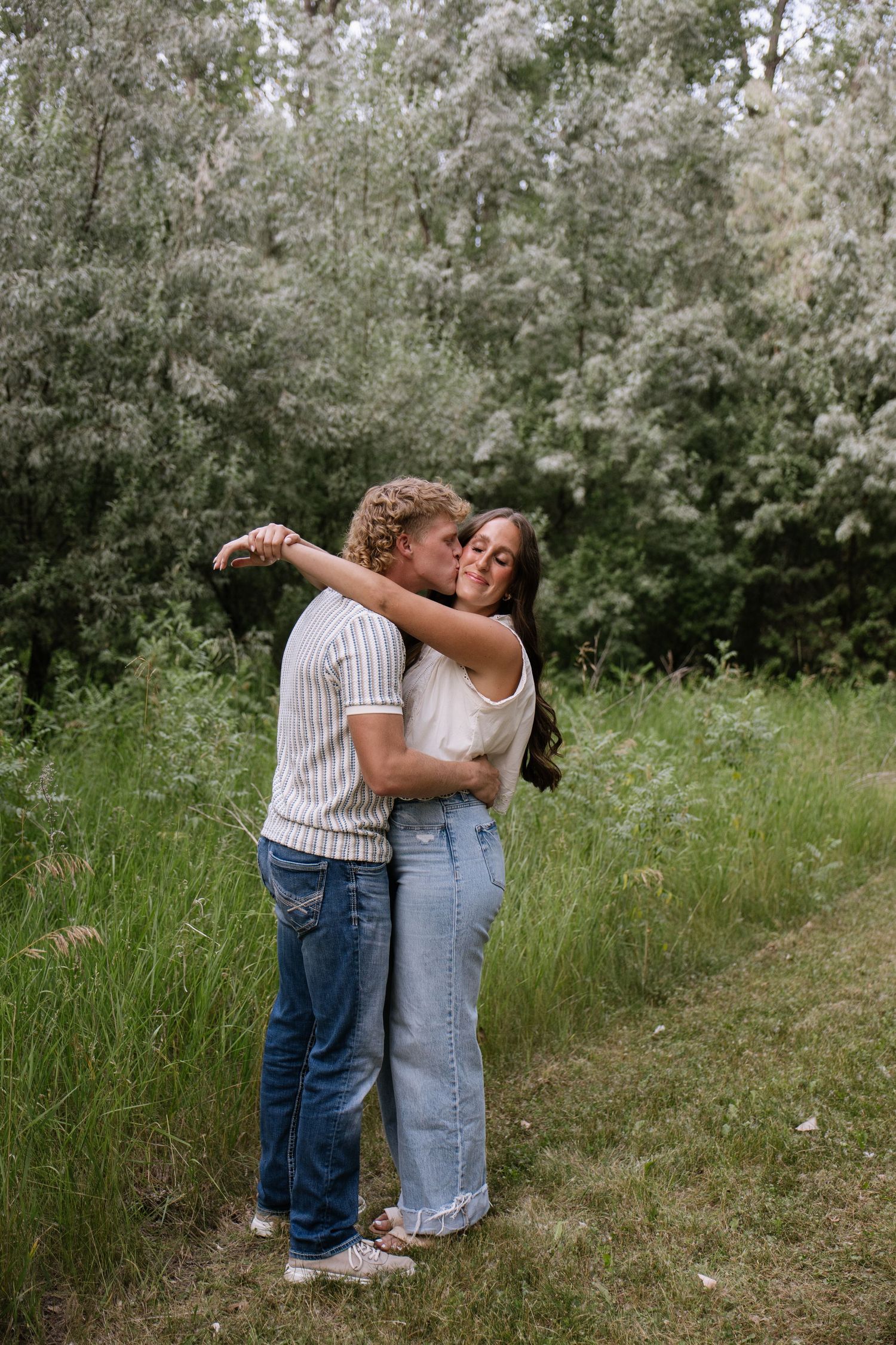 A couple embracing and kissing outdoors in a lush green field, both wearing jeans and casual tops.