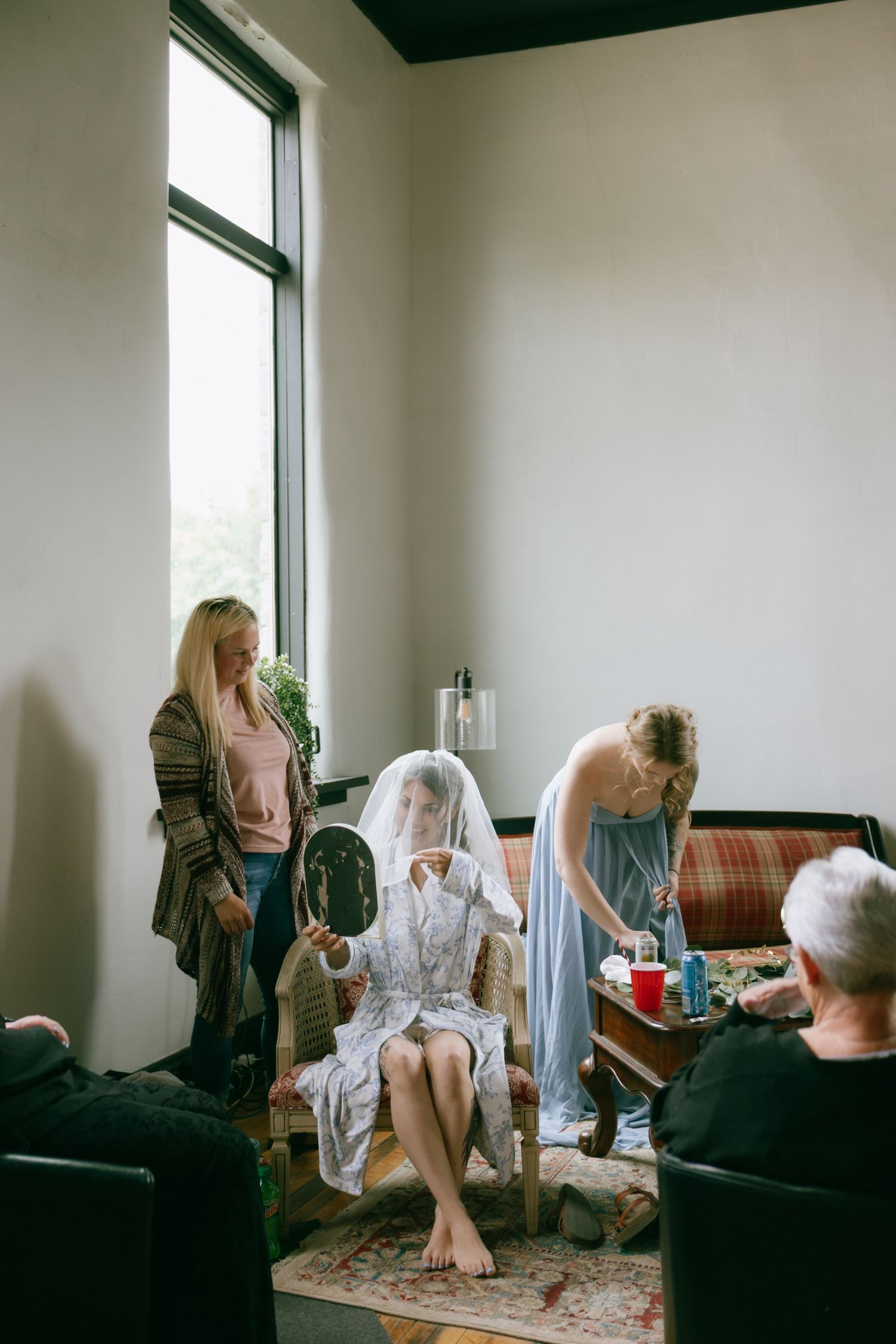 Bride in veil sits in chair holding mirror while bridesmaids assist her in a bright modern room.