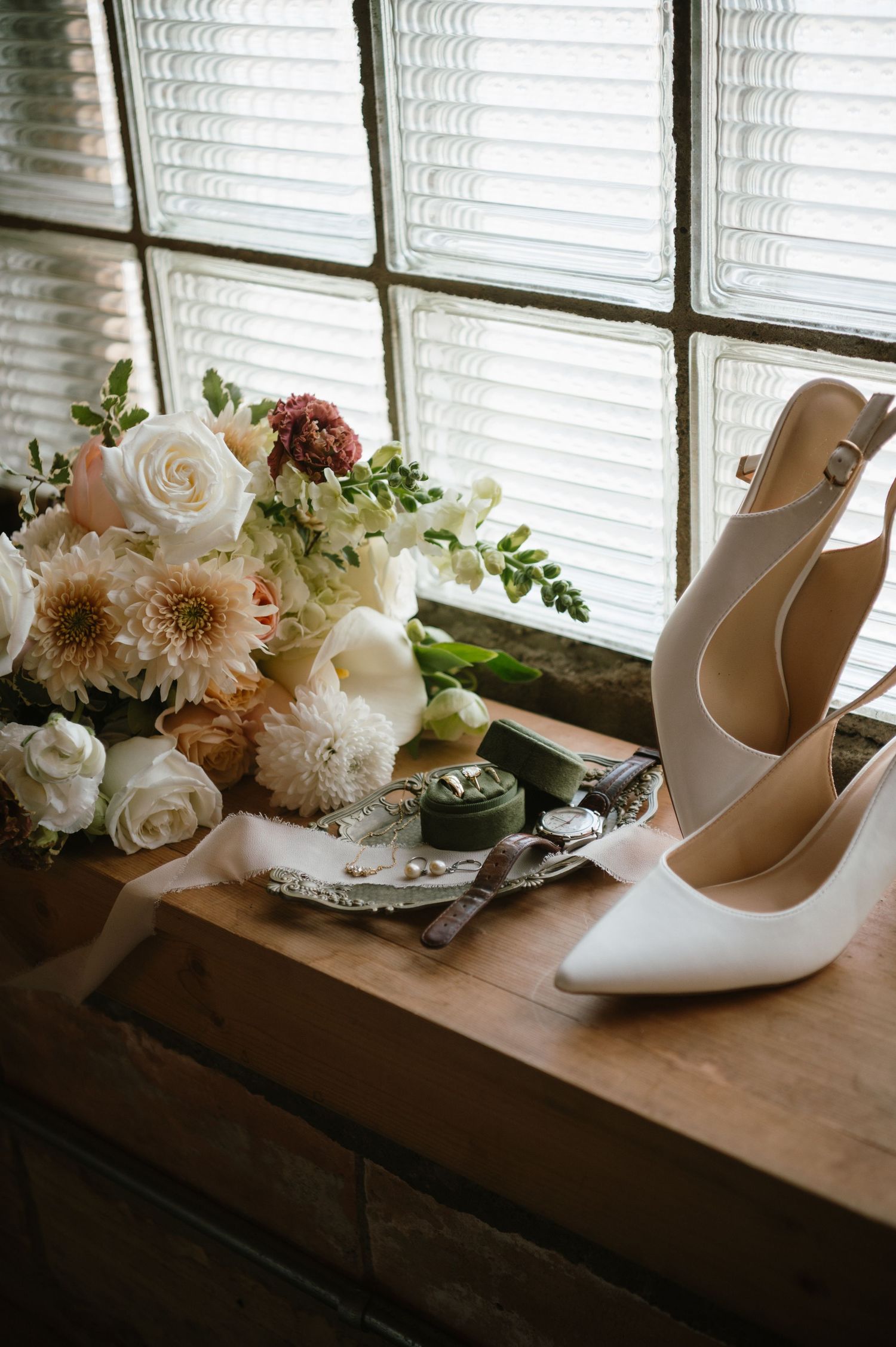 Elegant bridal flat lay with nude heels, white and blush floral bouquet, jewelry, and rings on a wooden table.