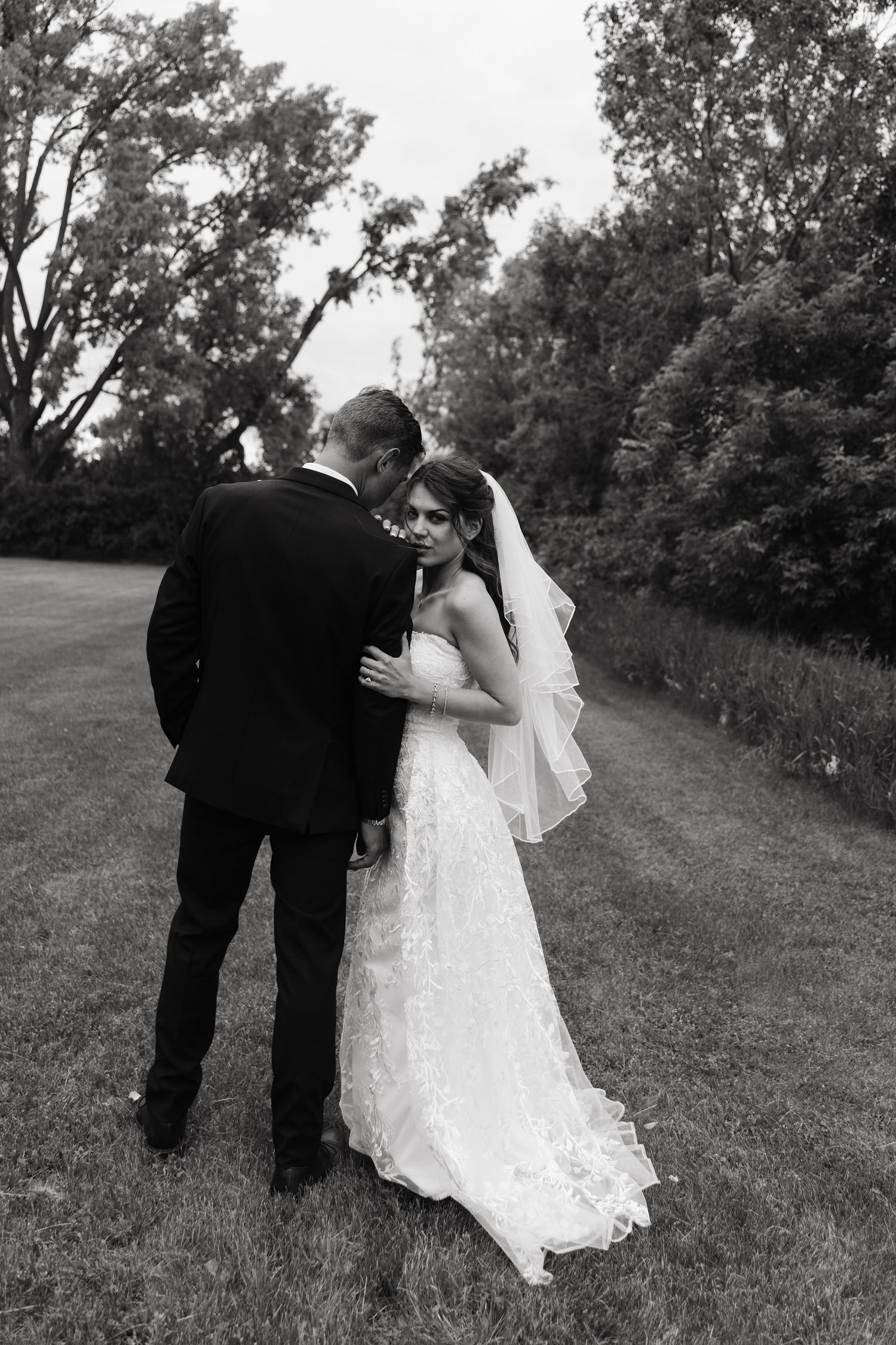 Black and white photo of a bride and groom embracing outdoors on a lush green lawn surrounded by trees.