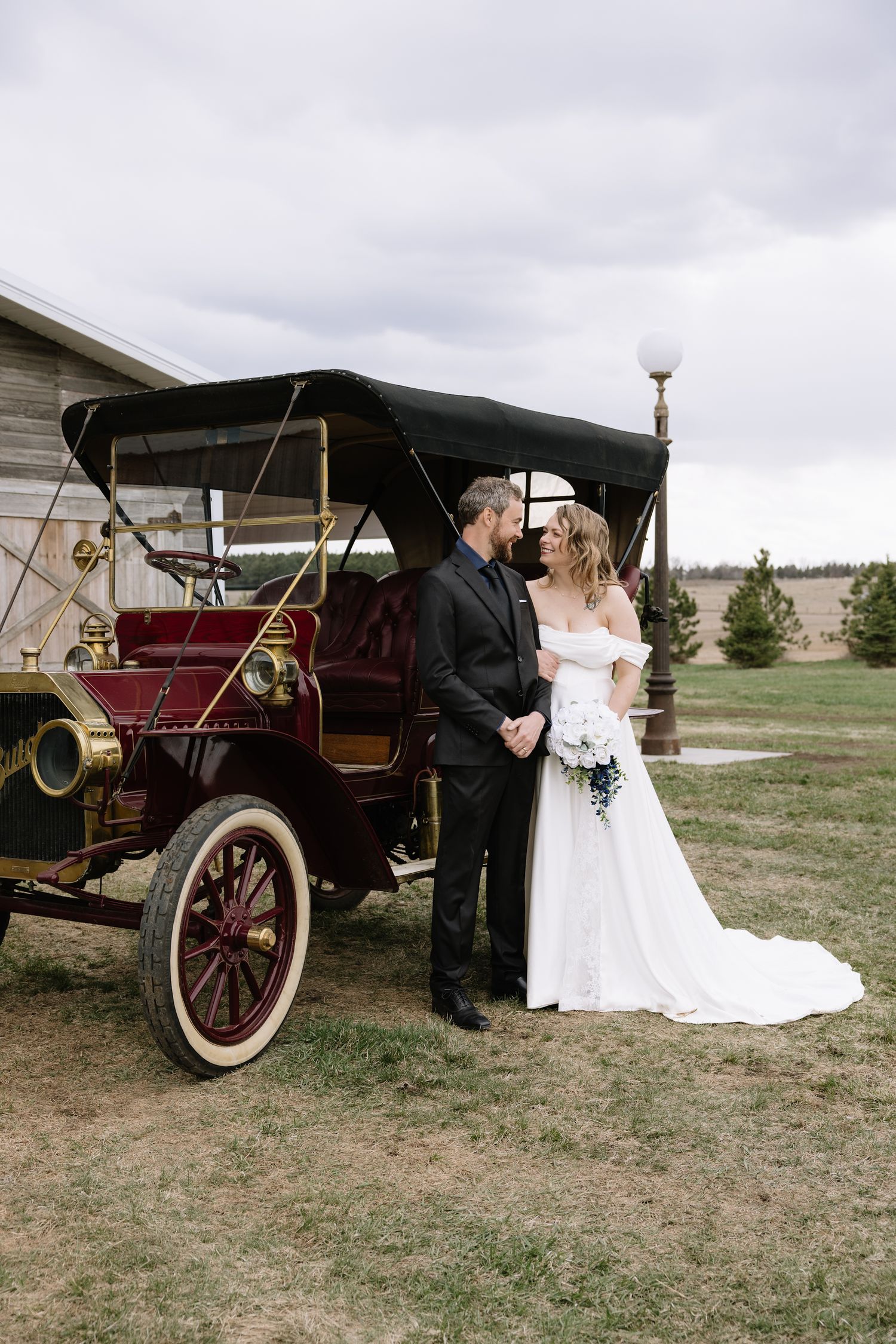 Bride and groom posing beside a vintage red antique car on a cloudy day at a rustic outdoor wedding venue.
