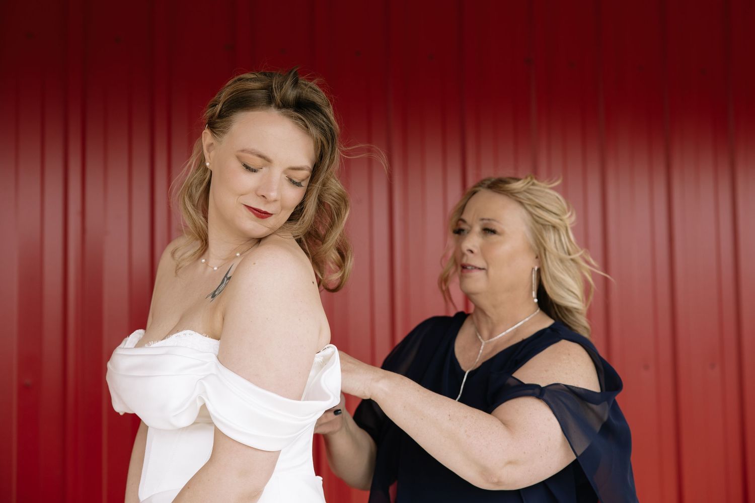 A mother helps her daughter button a white off-shoulder wedding dress in front of a red barn wall.