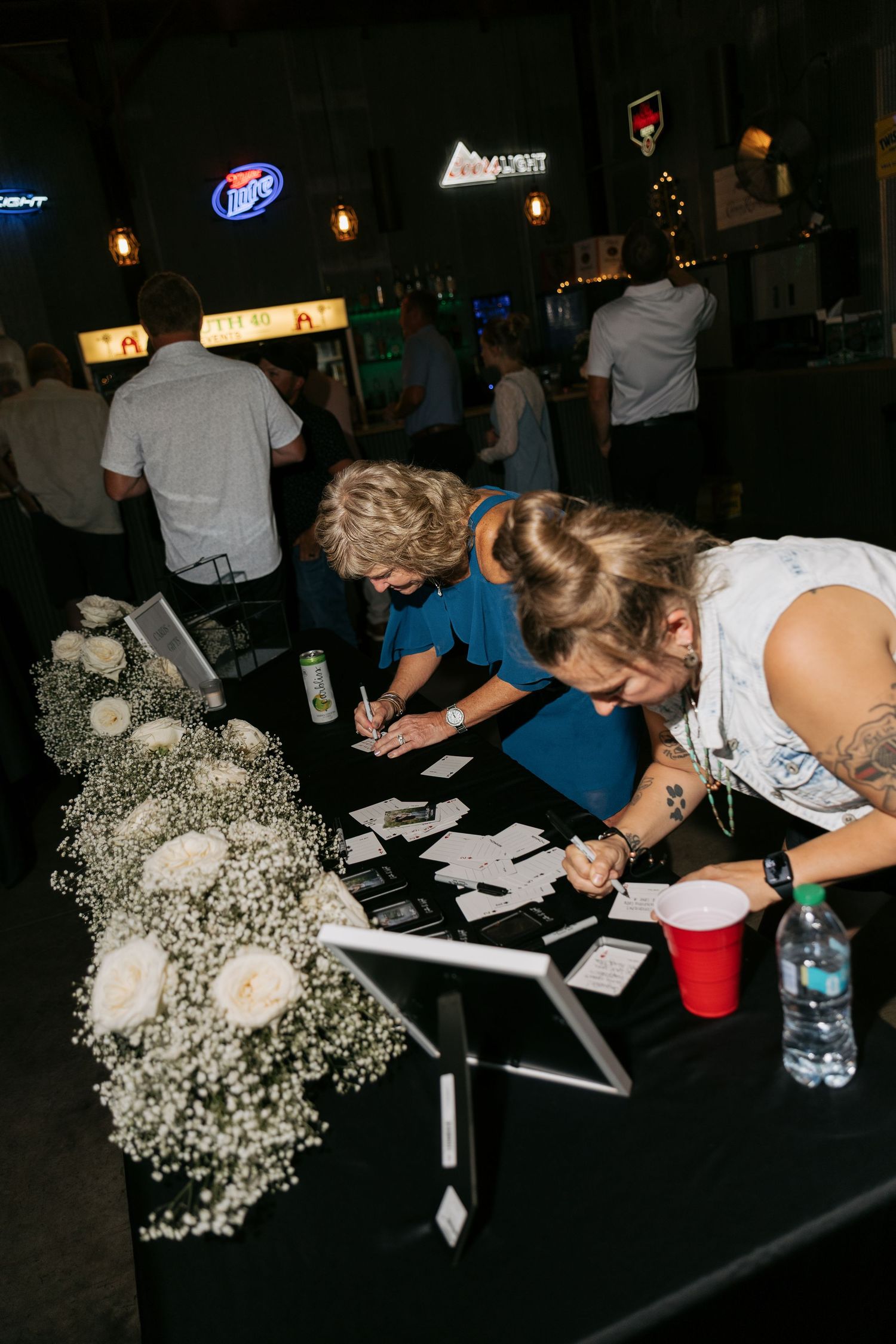 Two women signing documents at a decorated event registration table with white roses and baby's breath floral arrangements.