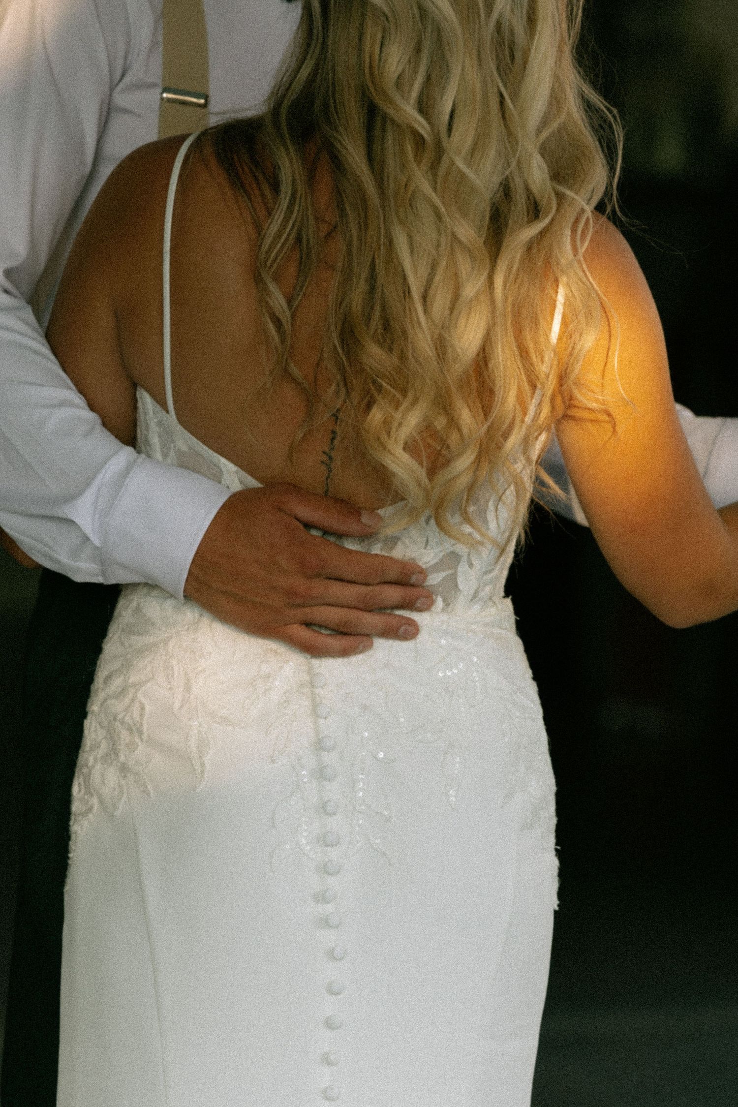 Bride in white lace wedding gown with button back detail, long blonde wavy hair, embraced by groom in white shirt.
