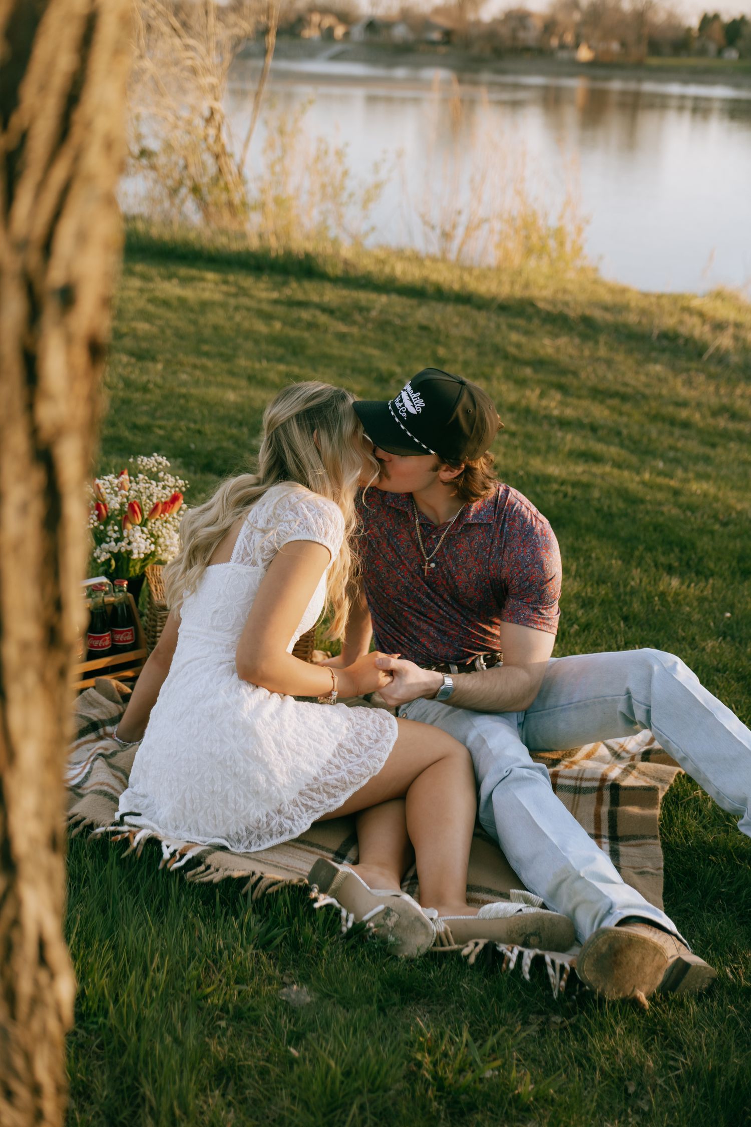 Couple sharing a tender kiss while sitting on a blanket by a serene lake during a golden hour picnic date.