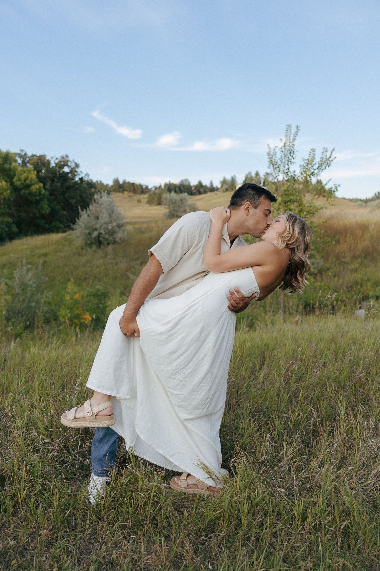 A couple shares a romantic dip kiss in a sunlit meadow with rolling hills in the background.