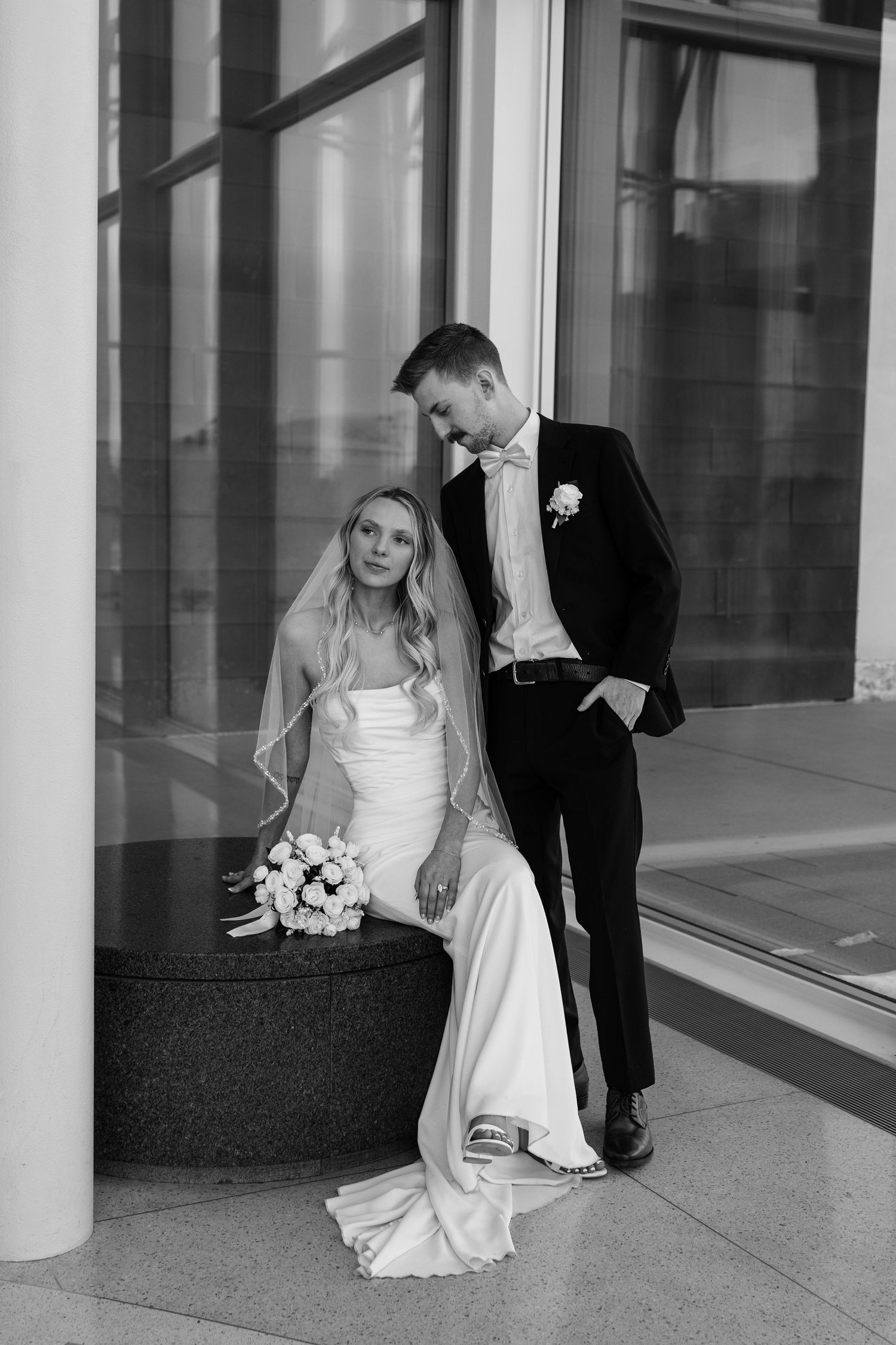 A bride and groom share a romantic moment between elegant white columns in a modern building.