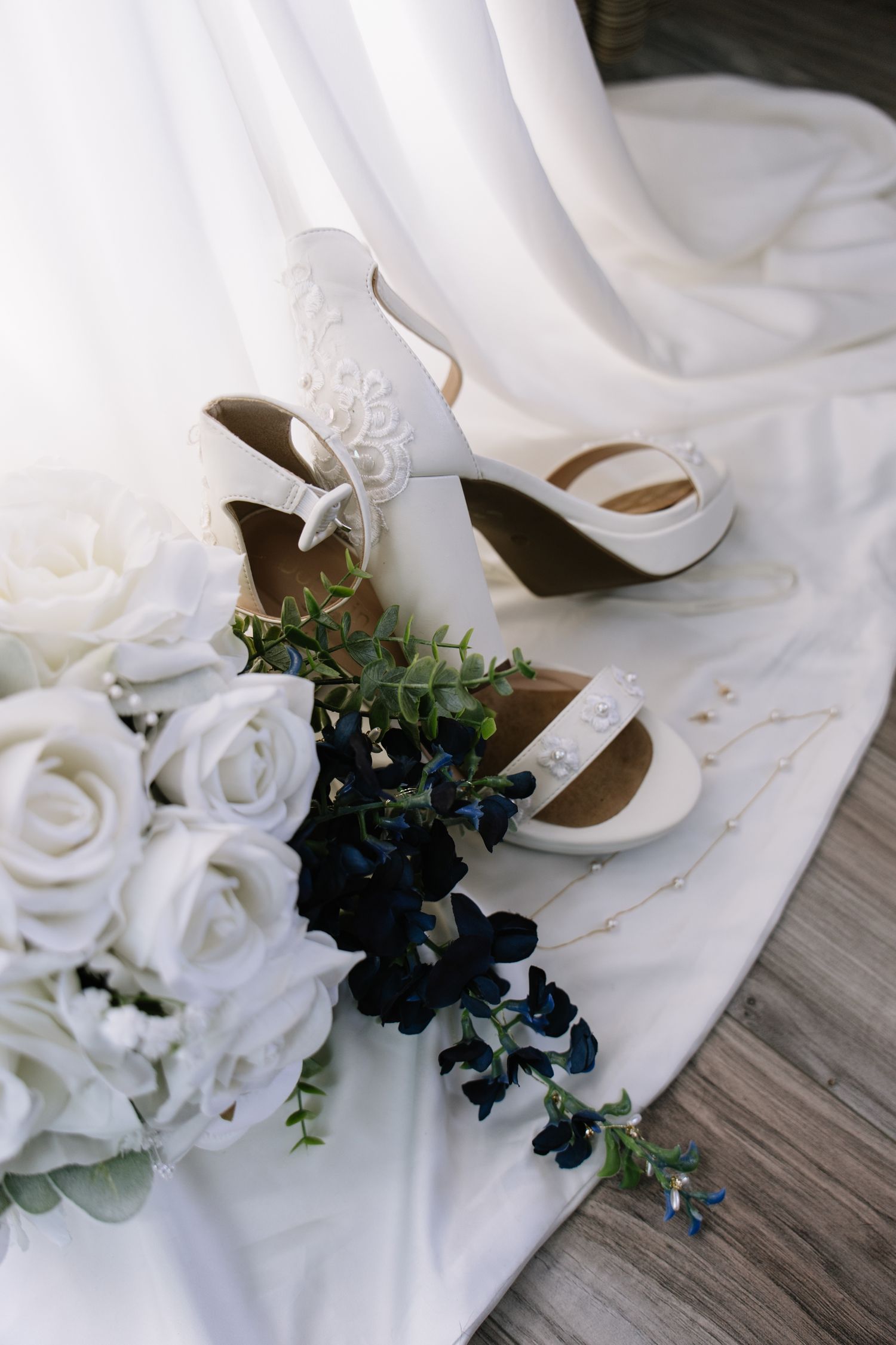 Bridal white heeled shoes with lace detail beside a bouquet of white roses and navy blue flowers on a white dress.