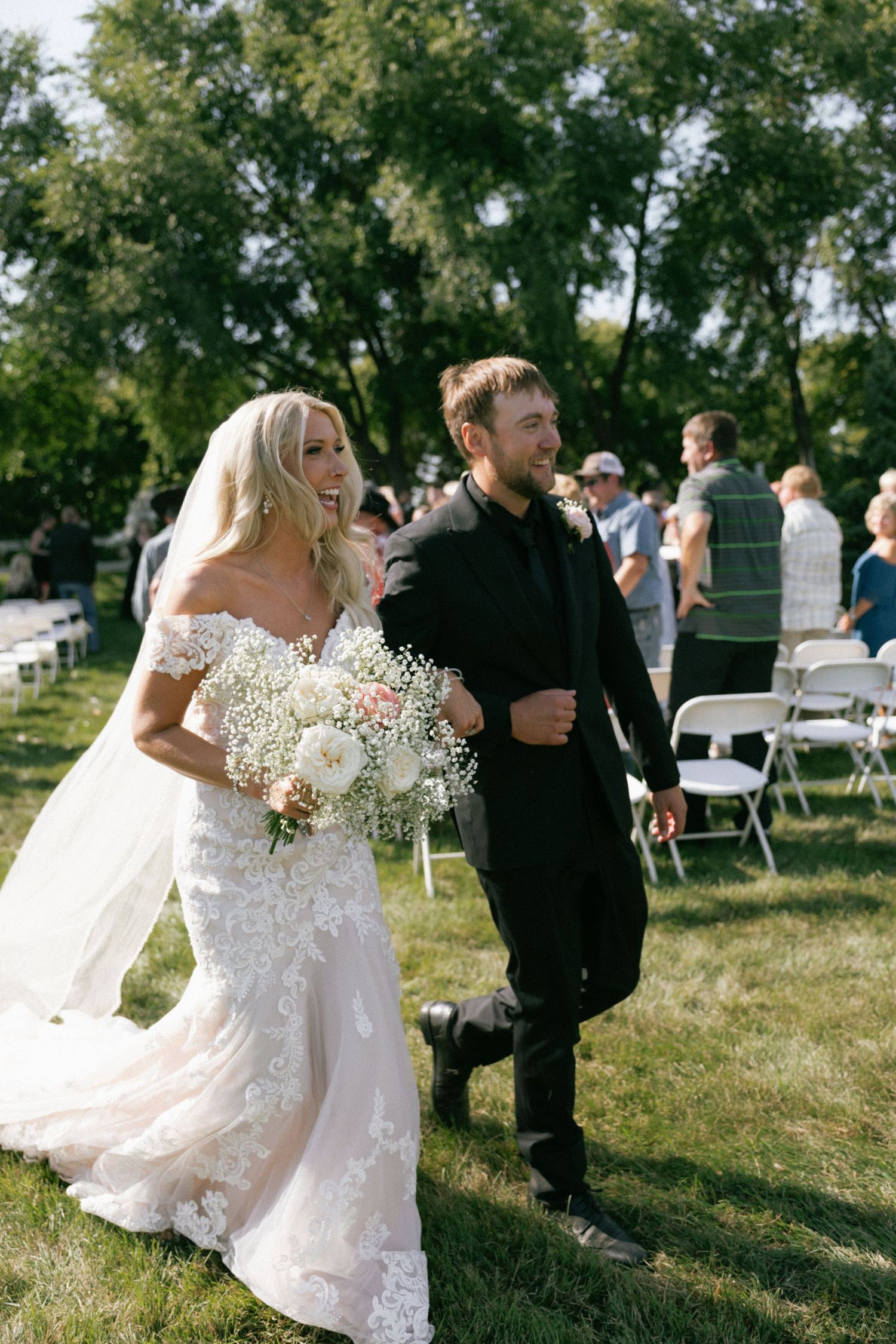 Bride in lace gown holding white bouquet walks with groom in black suit down outdoor wedding aisle on sunny day.