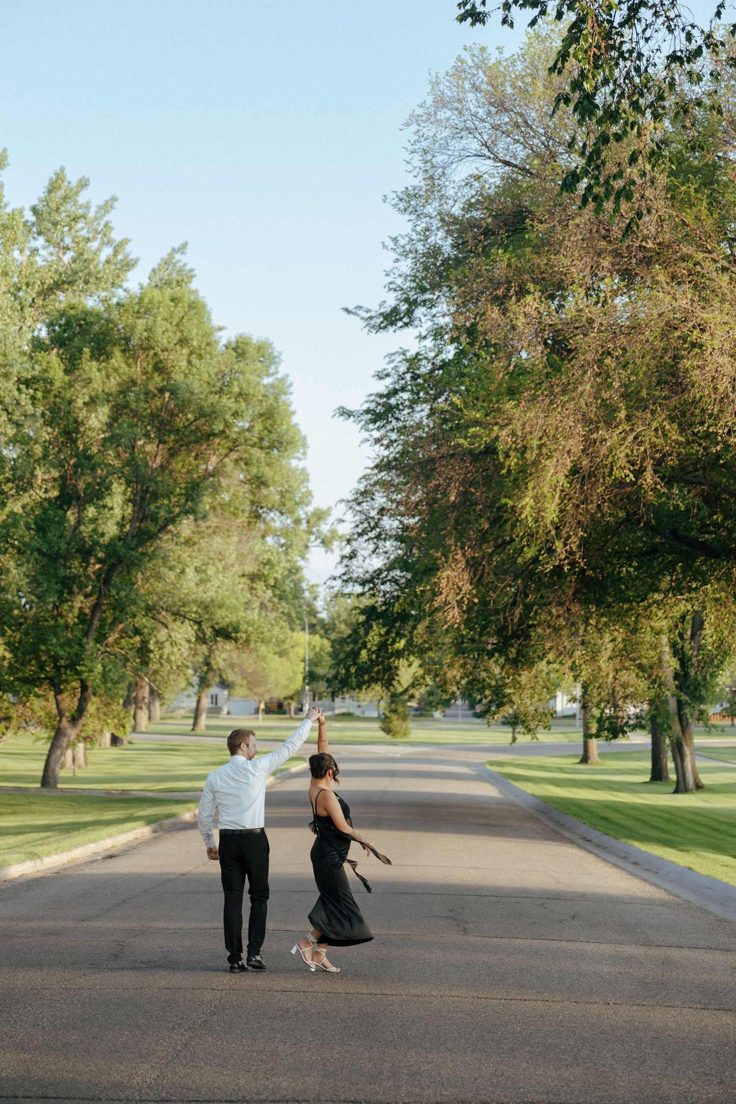 A couple dances playfully together on a tree-lined path in a park setting.