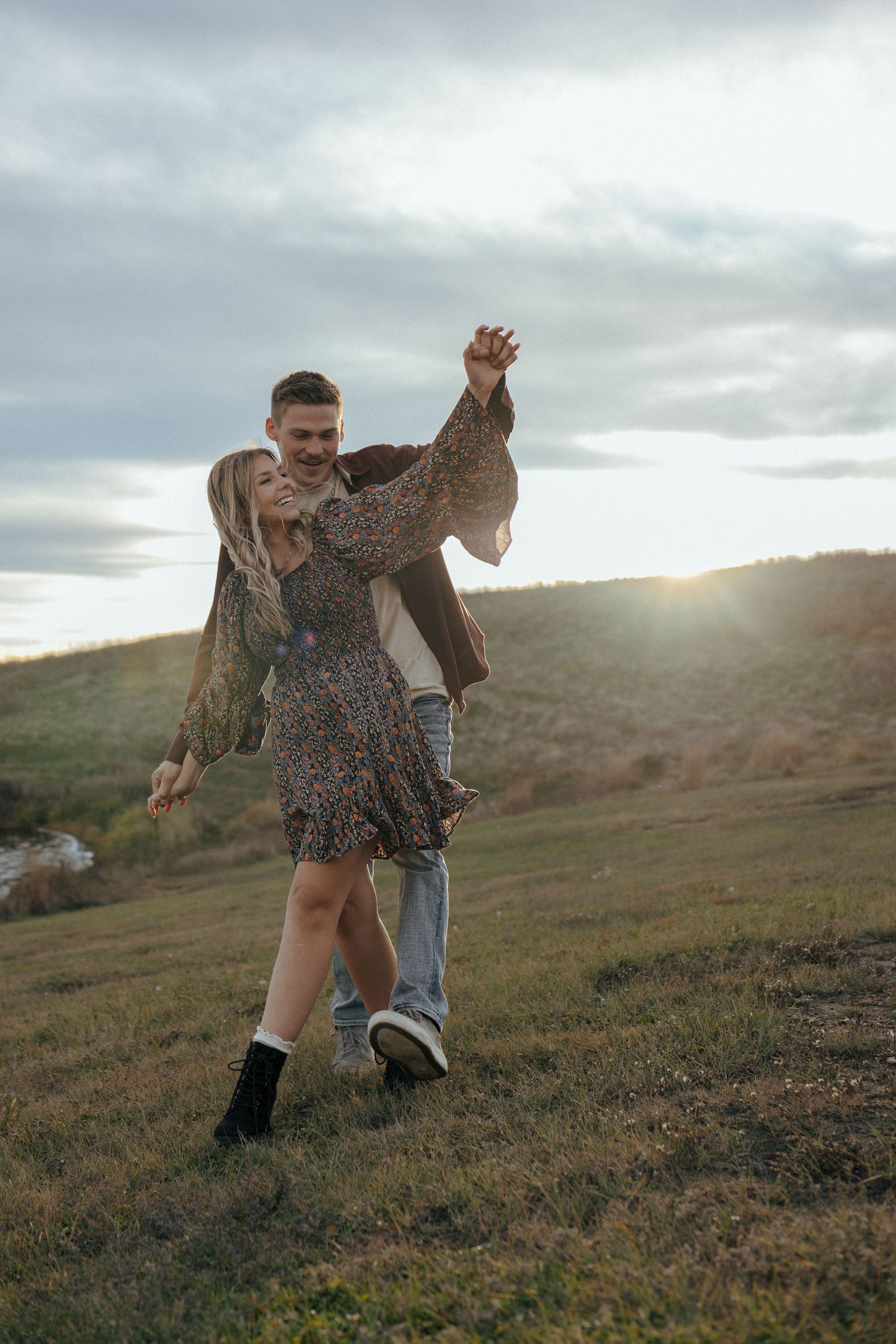 Playful couple dancing and twirling on a hillside at dusk.