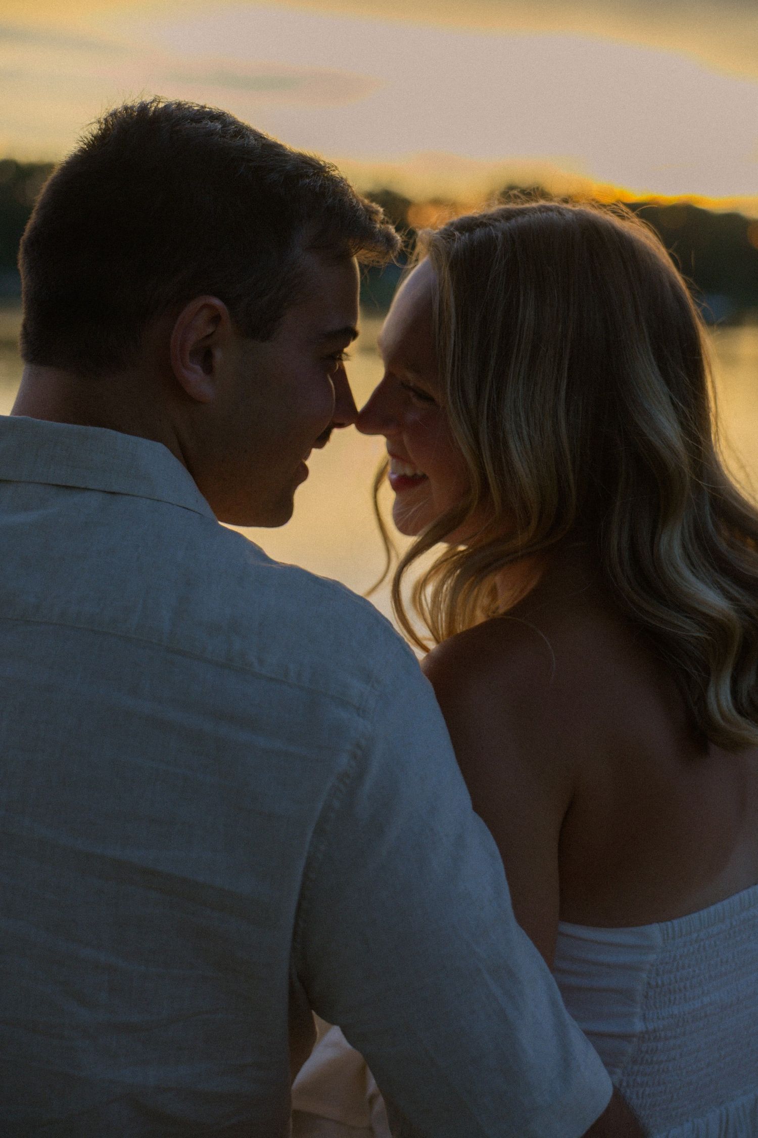 Silhouettes of a couple sharing tender moments against a sunset sky by the water.