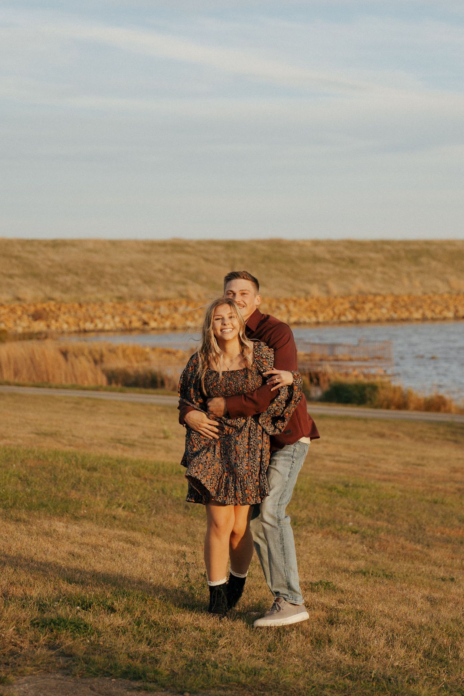 A couple shares a joyful moment on a grassy hillside overlooking a lake.