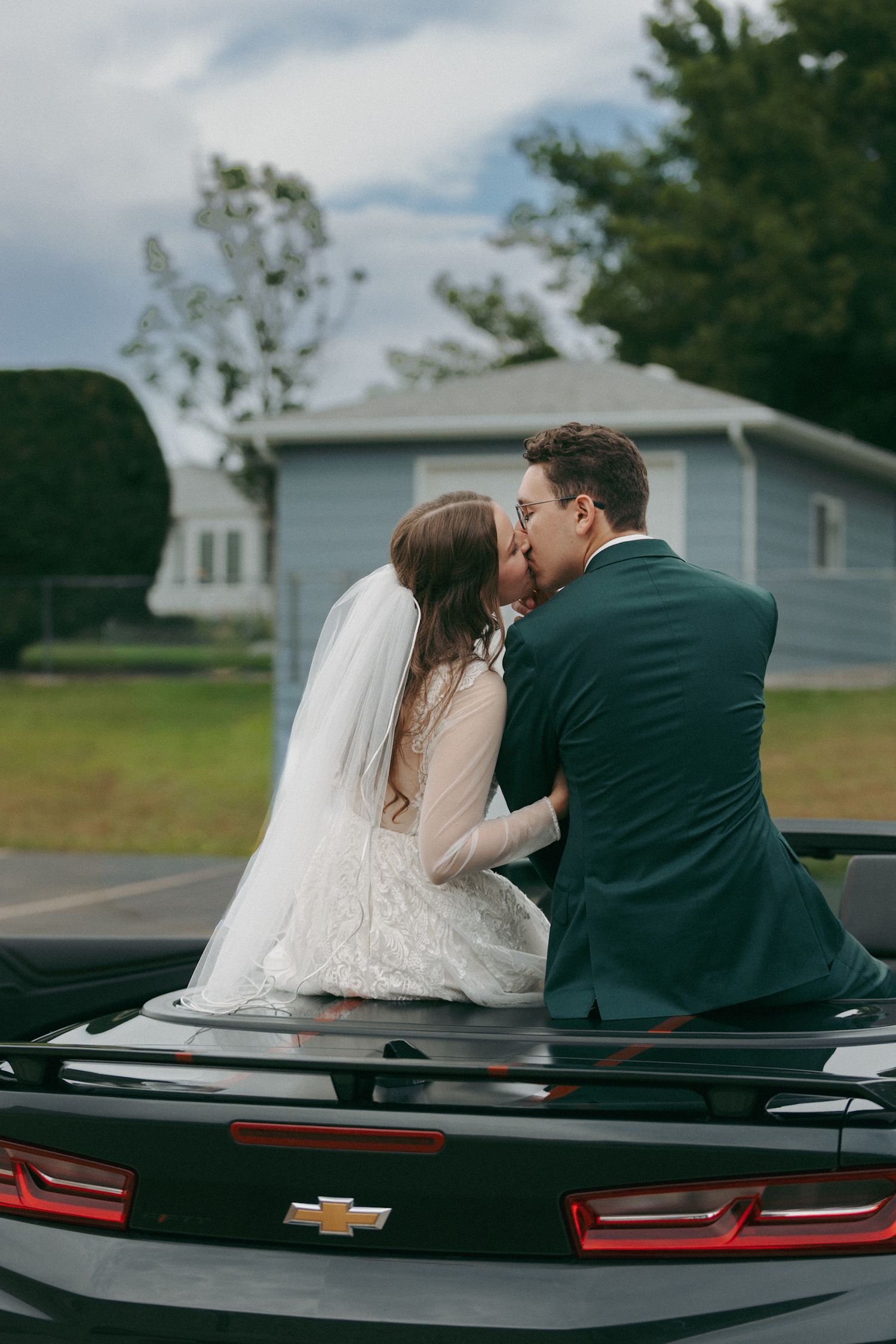 Wedding couple sharing a kiss while sitting on the trunk of a black Chevrolet car.