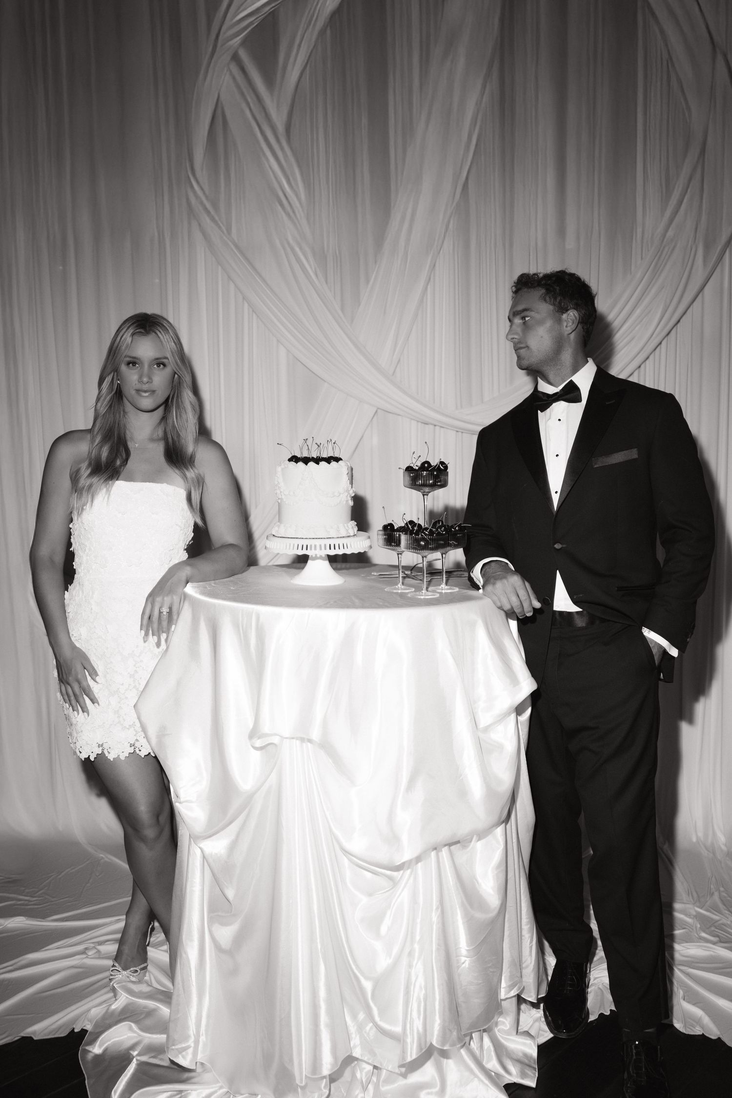 Black and white photo of a bride and groom standing beside a wedding cake at their elegant reception.