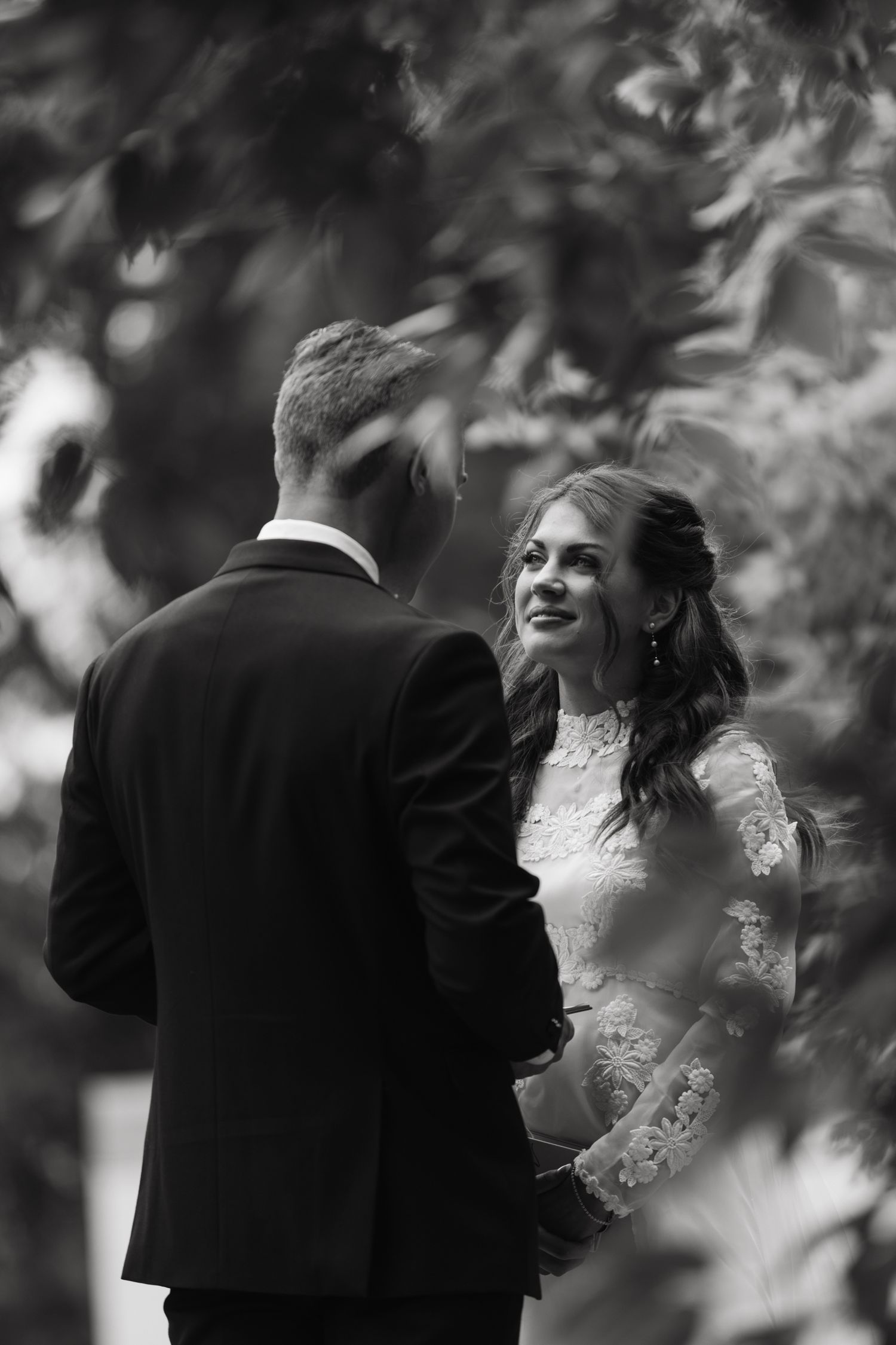 Black and white photo of a smiling bride in lace gown holding hands with groom in suit outdoors.