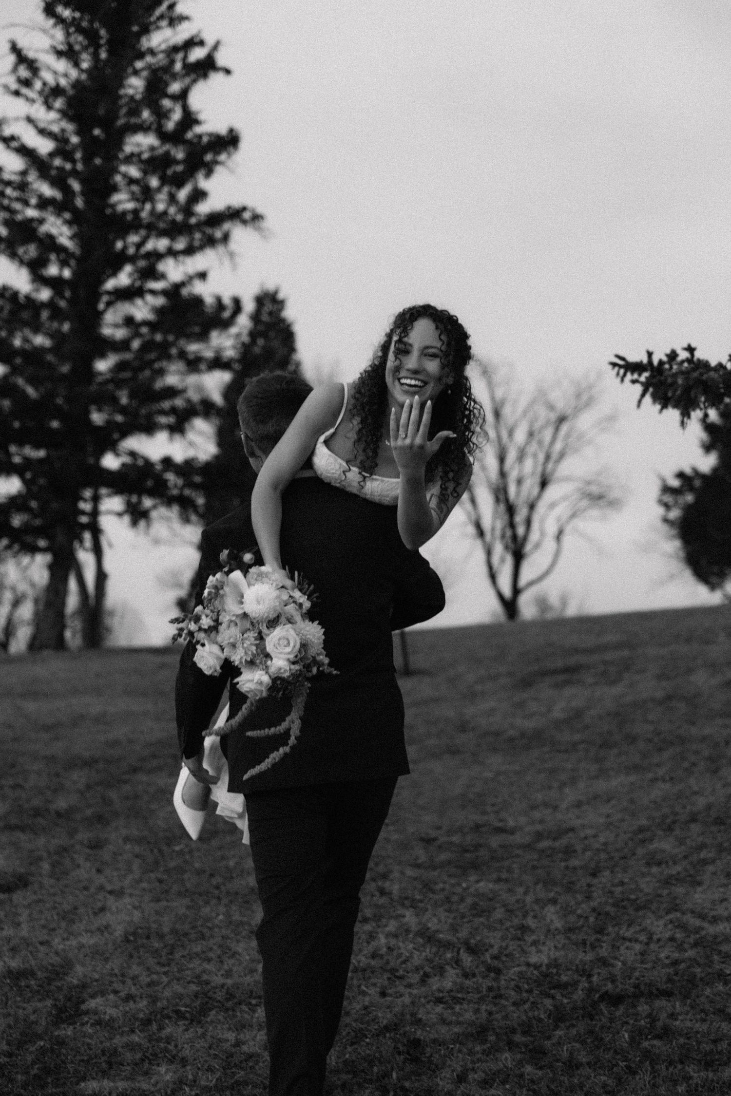 Black and white photo of groom lifting smiling bride holding bouquet outdoors near trees.