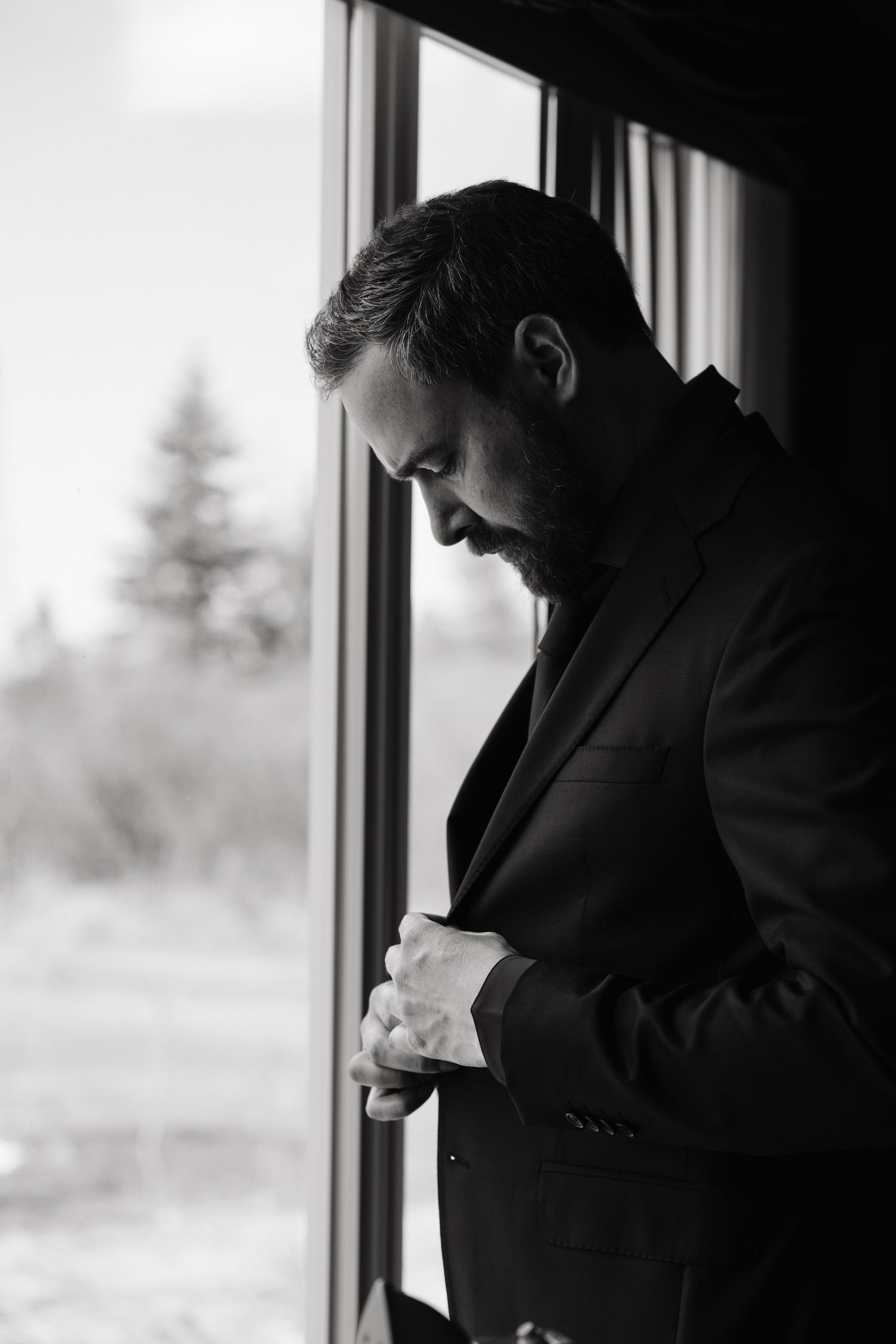 Black and white portrait of a bearded man in a suit adjusting his jacket by a window with trees outside.