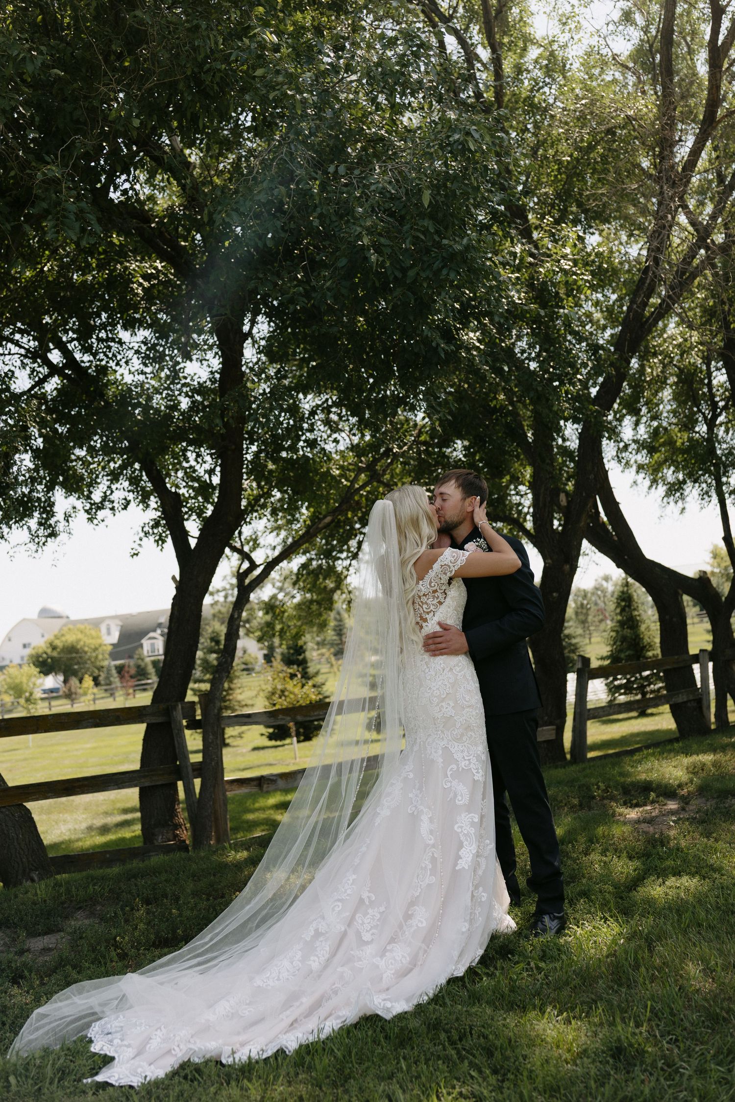 Bride and groom share a romantic kiss outdoors, surrounded by lush green trees, with a flowing lace wedding veil trailing on the grass.