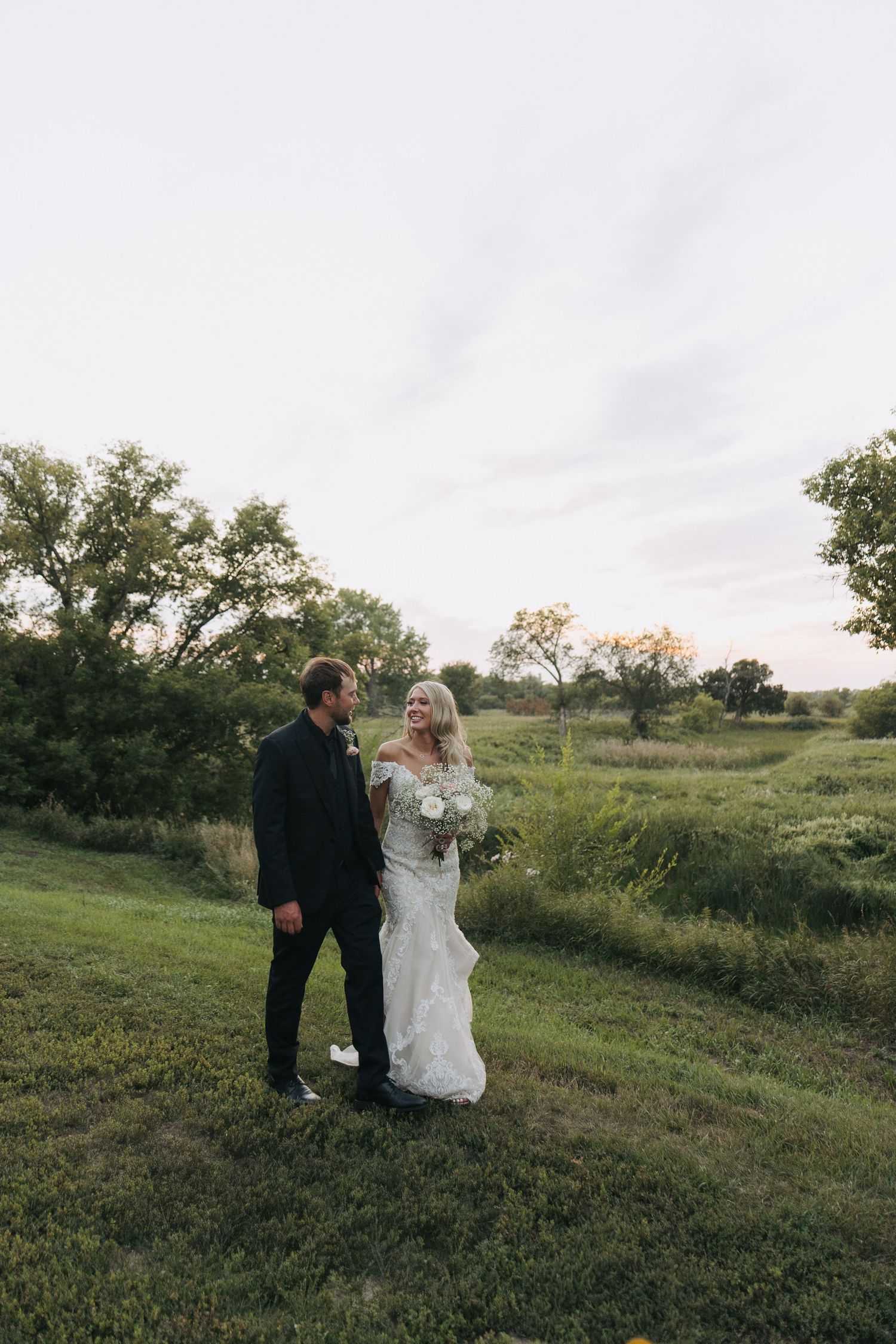 Bride in lace gown and groom in black suit pose on green hillside at sunset wedding.