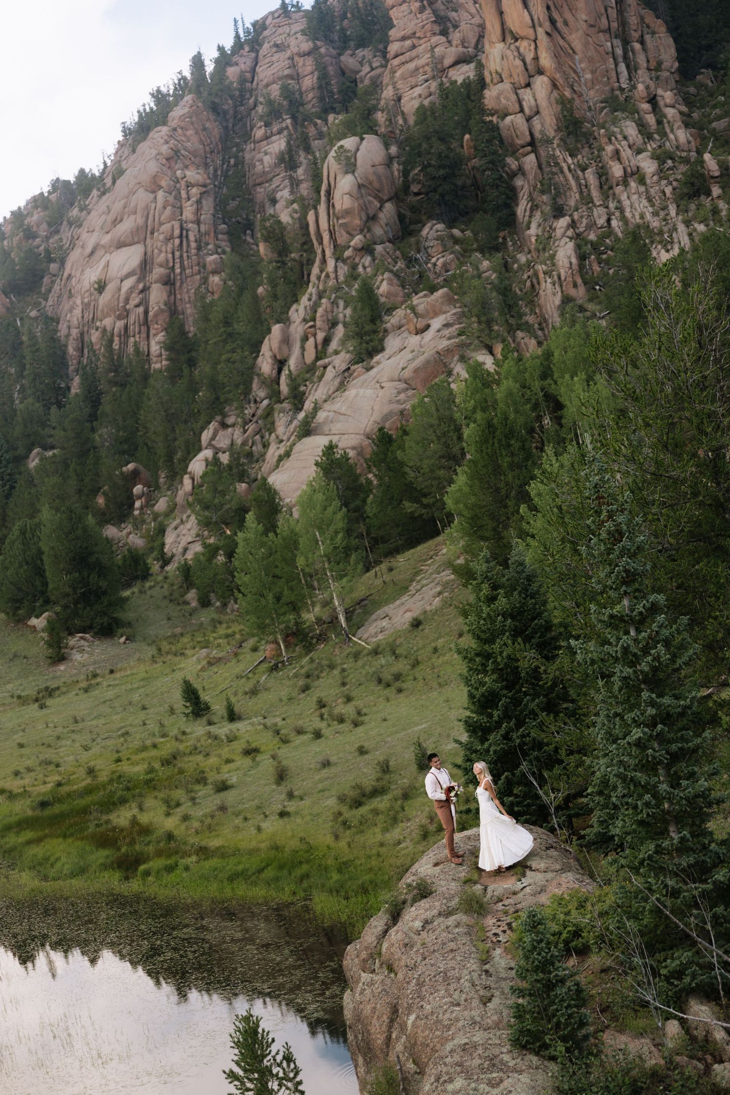 Bride and groom stand on rocky cliff beside mountain lake surrounded by pine trees and dramatic stone cliffs.