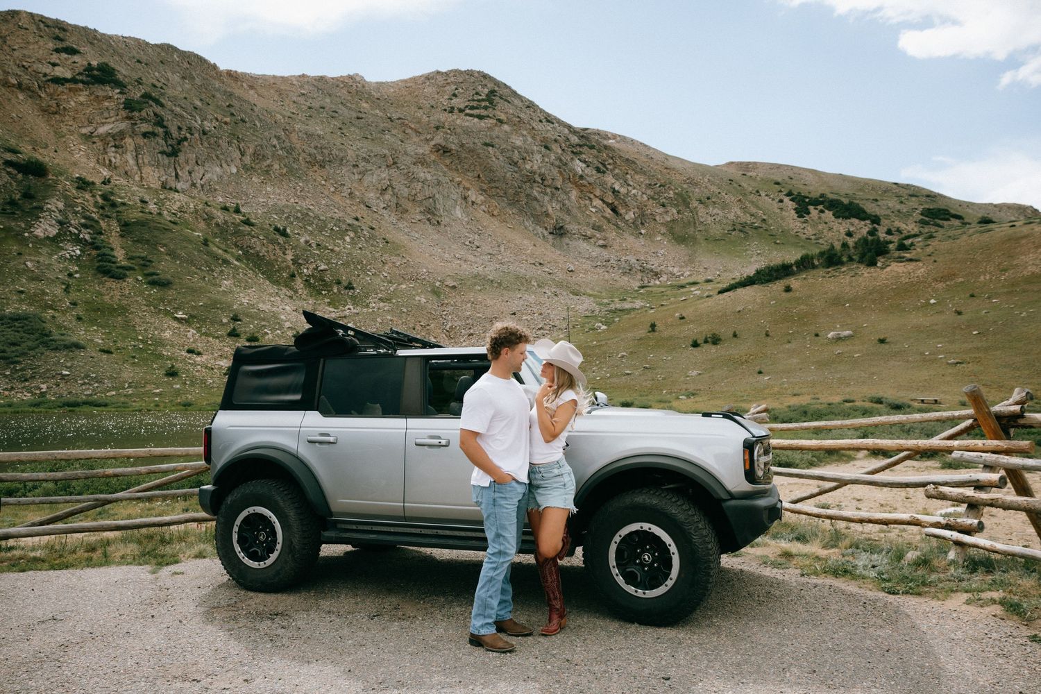 Couple kissing beside a silver Ford Bronco on a mountain road with scenic rocky hills in the background.