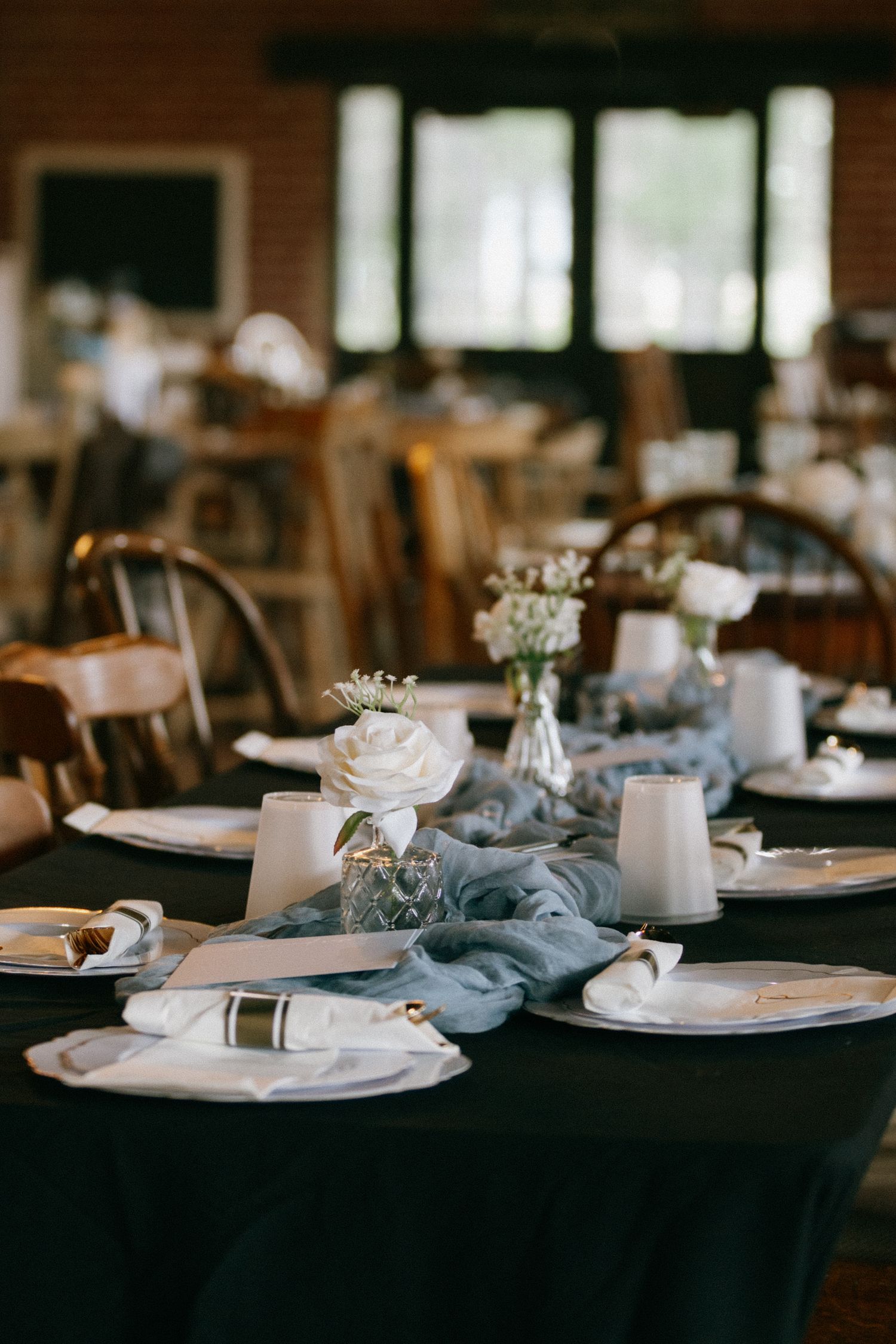 Elegant wedding reception table with dusty blue runner, white roses, candles, and white place settings on a dark tablecloth.