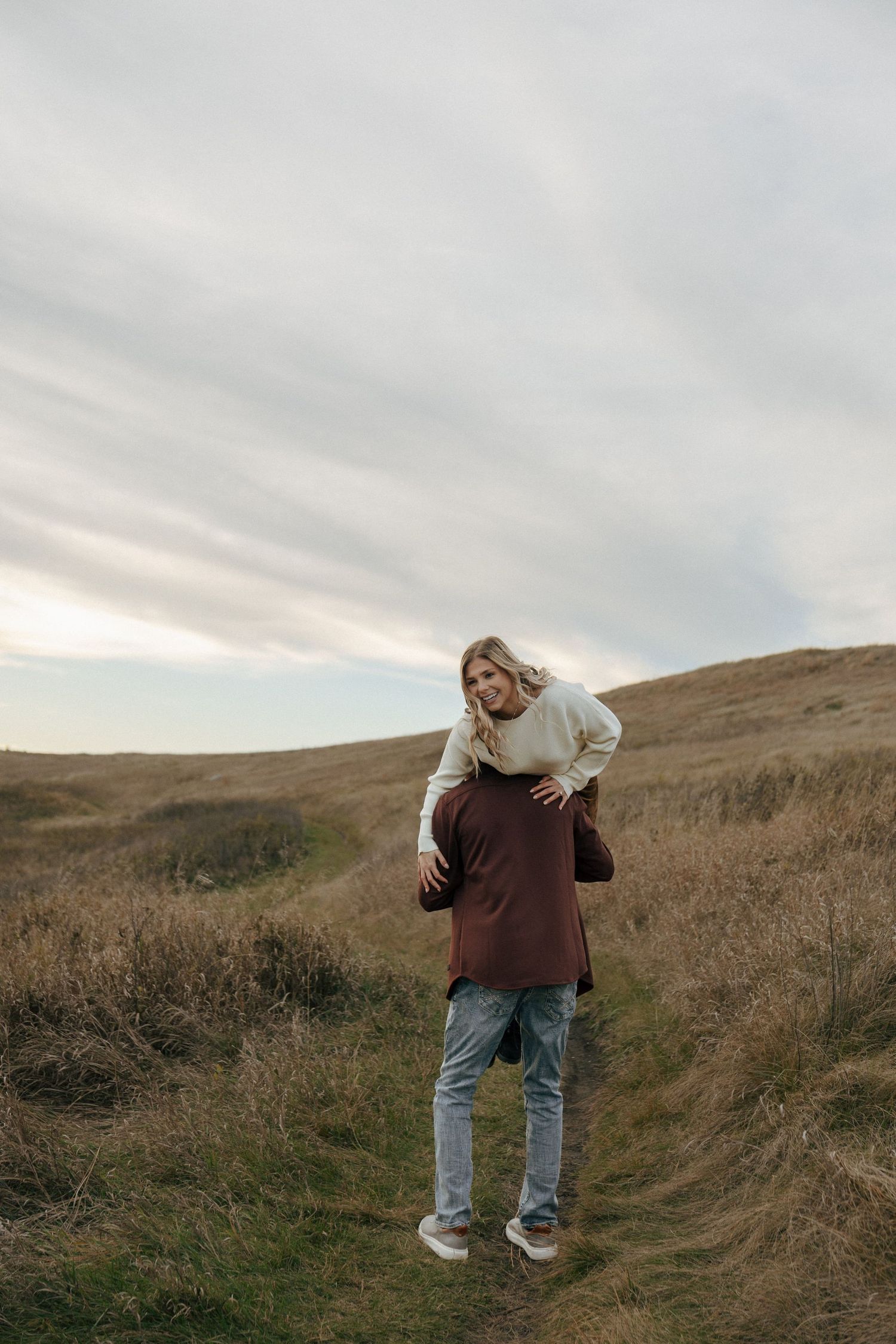 A person in jeans and sweater stands playfully on a grassy hillside path.