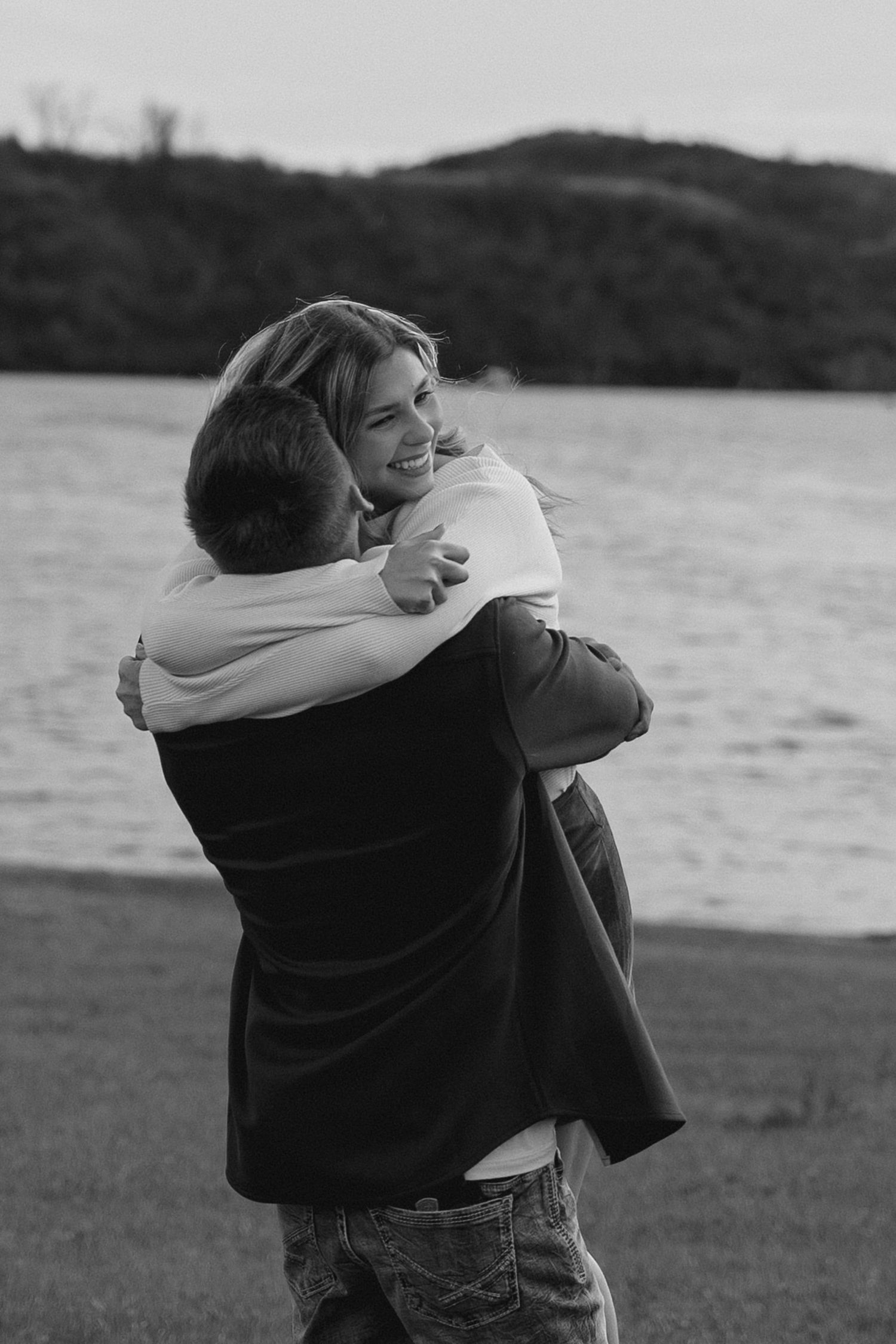 A black and white photo shows two people hugging by a lake with mountains in the background.