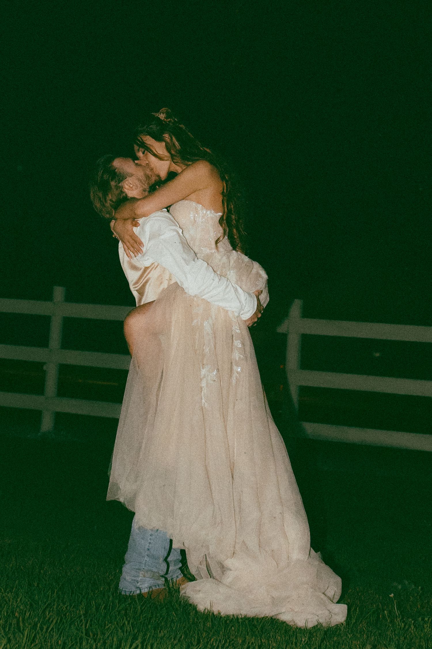 Romantic nighttime wedding photos with couple embracing on grass near white fence.
