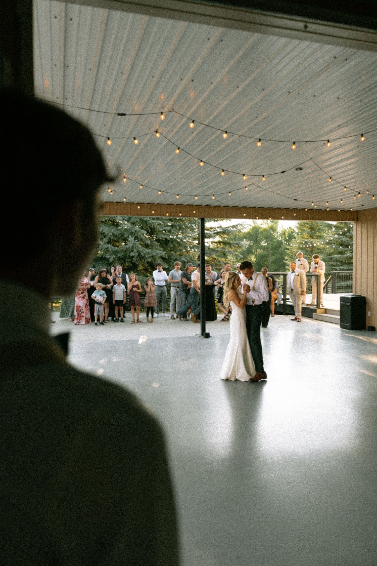 Bride and groom share their first dance under string lights at an outdoor wedding reception with guests watching.