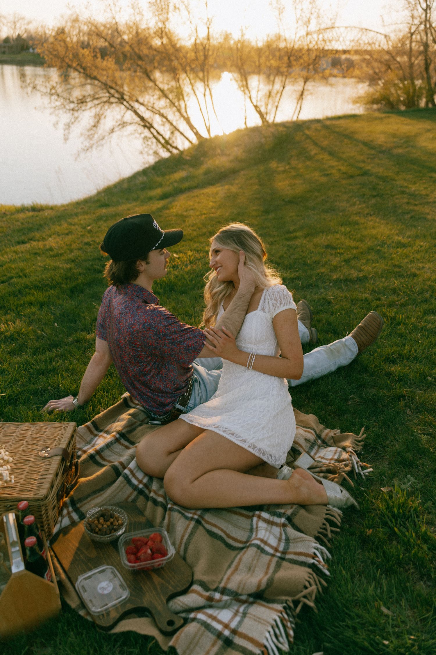 Couple sharing a romantic sunset picnic on a plaid blanket by the river, with strawberries and a wicker basket.