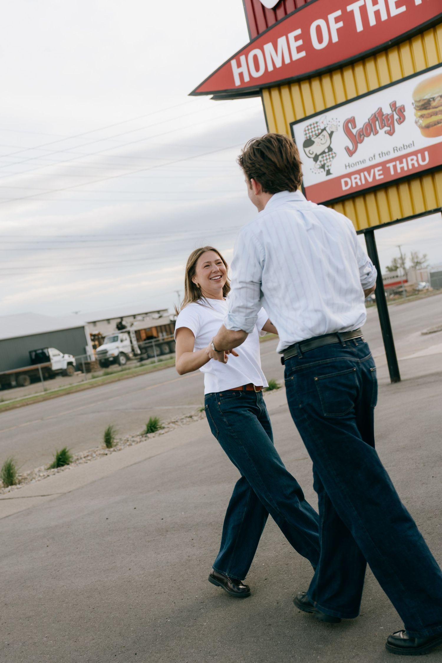 A laughing couple holds hands and dances in a parking lot near a Scotty's Drive Thru restaurant sign.