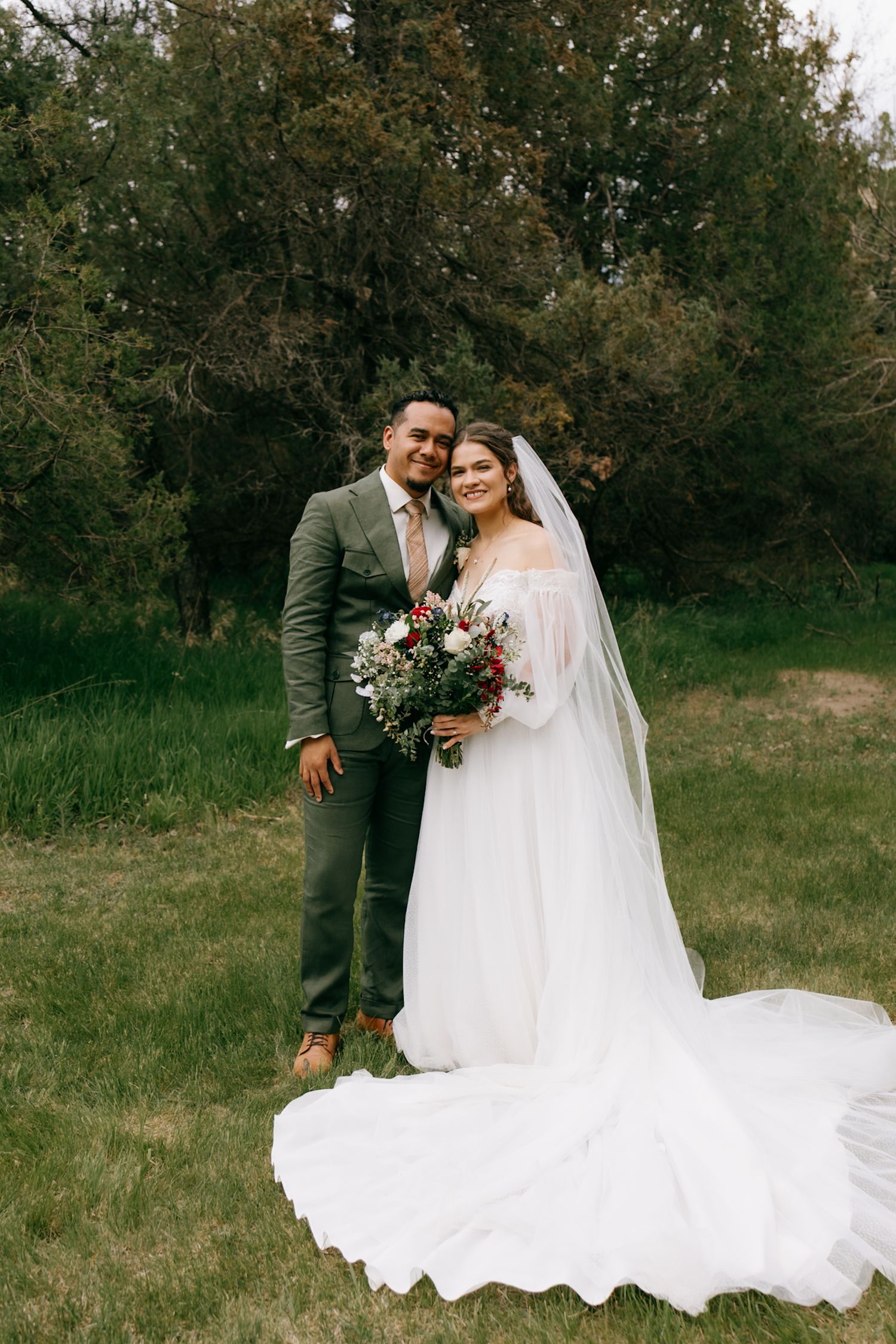 Bride in white gown with veil holding bouquet stands with groom in gray suit outdoors on green grass.