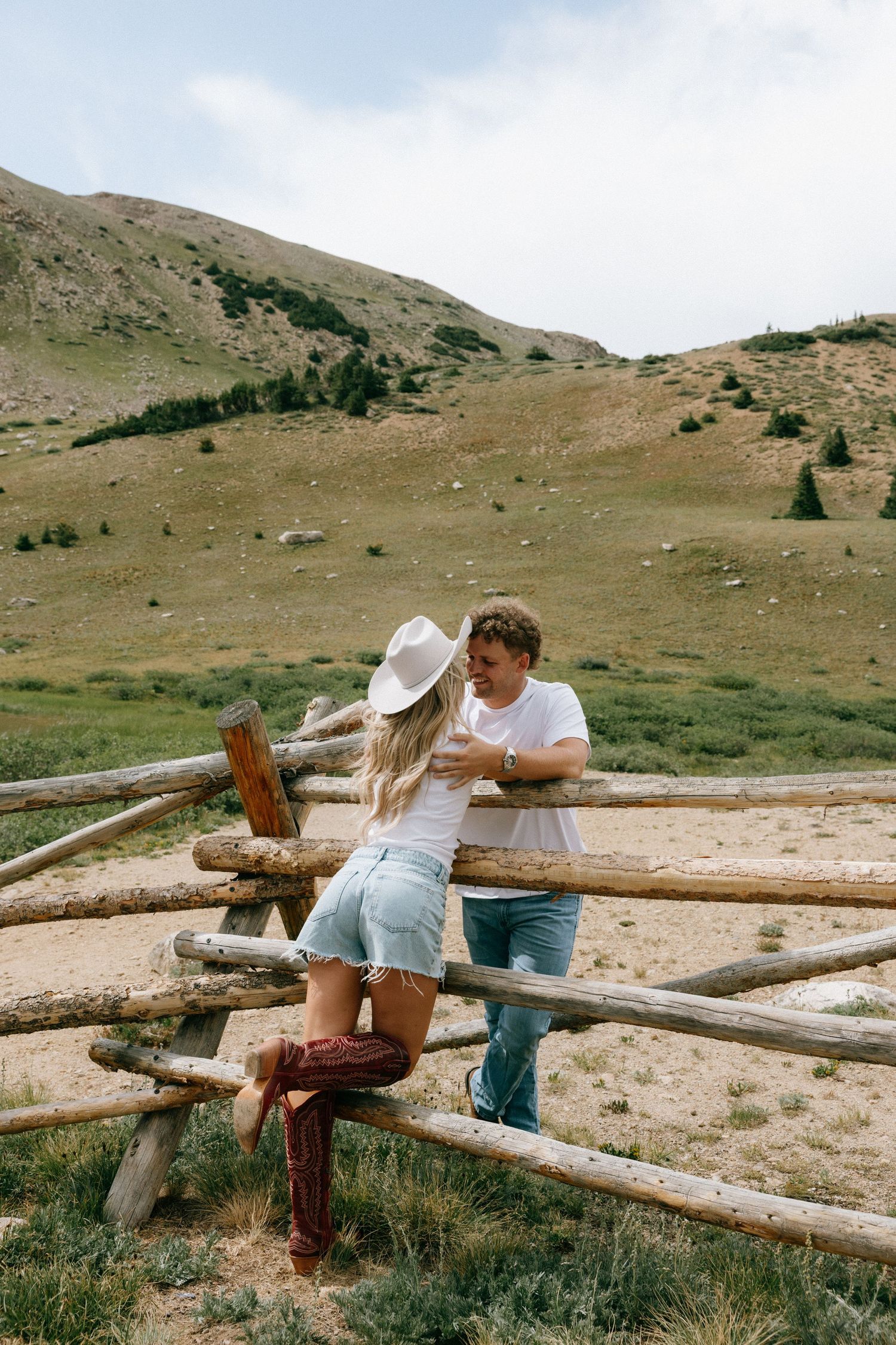 Couple leaning against rustic wooden fence in mountain meadow, woman wearing cowboy hat and boots, man in white tee.