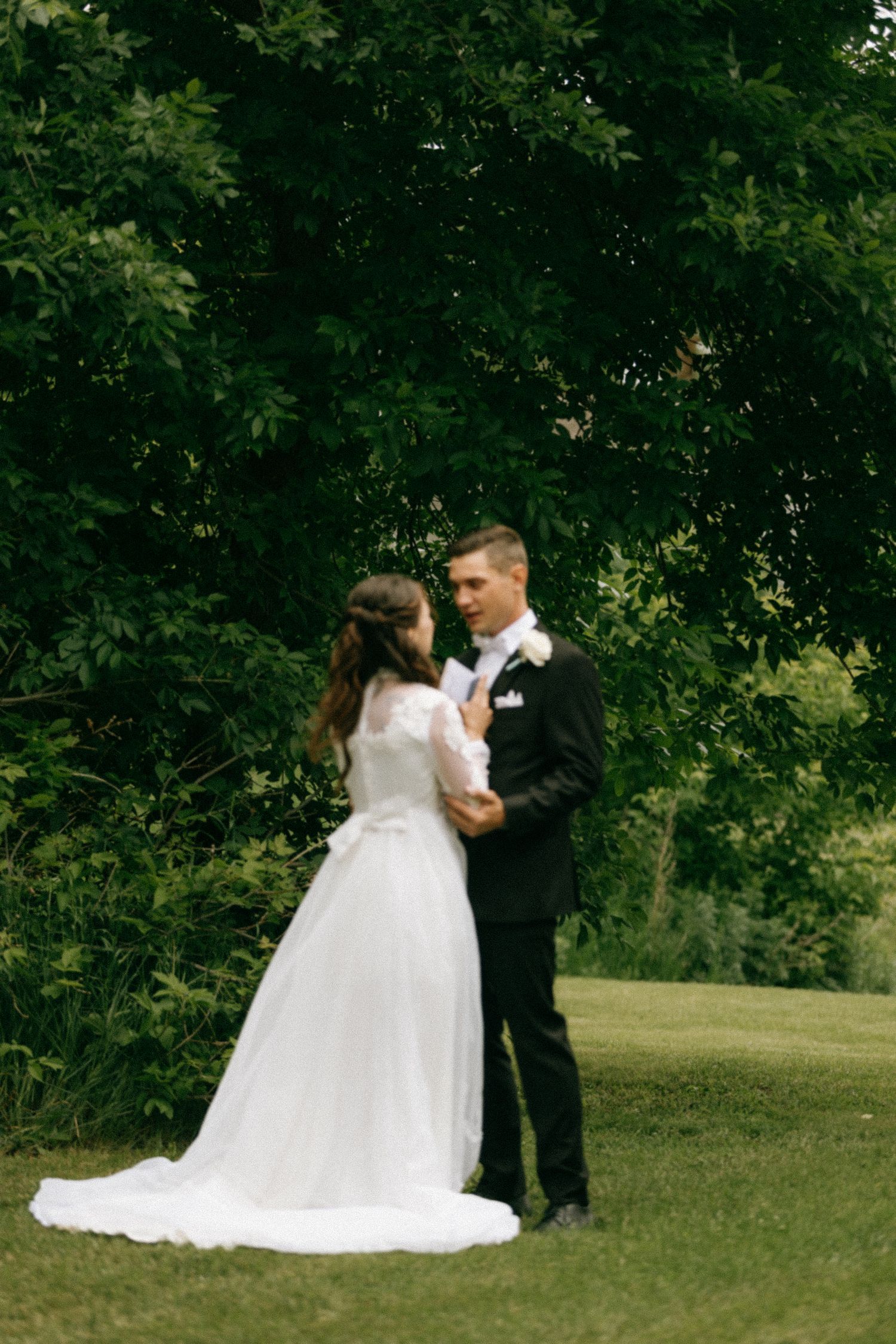 A bride in a white gown and groom in a black suit share a romantic moment on a green lawn surrounded by lush trees.