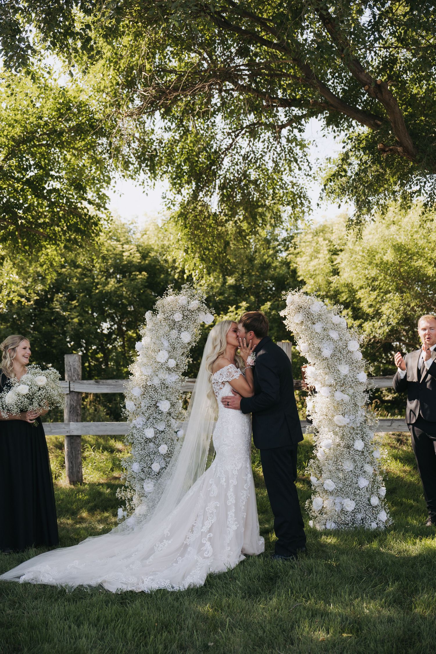 Bride and groom share first kiss under a stunning white floral arch at an outdoor garden wedding ceremony.