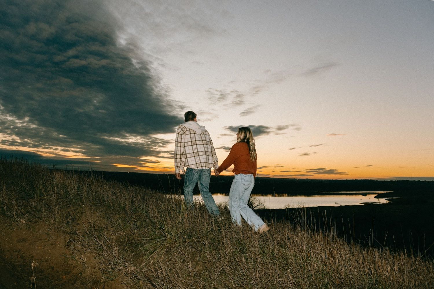 Silhouetted couple walking through grass field at sunset near water.