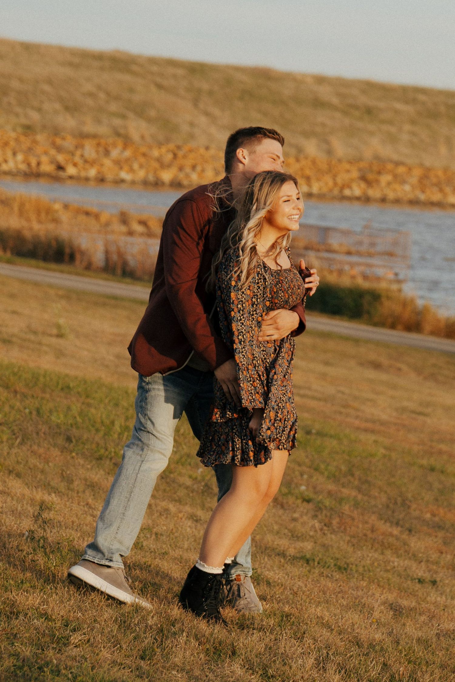 Two people share a playful moment on grass near a lake during sunset.