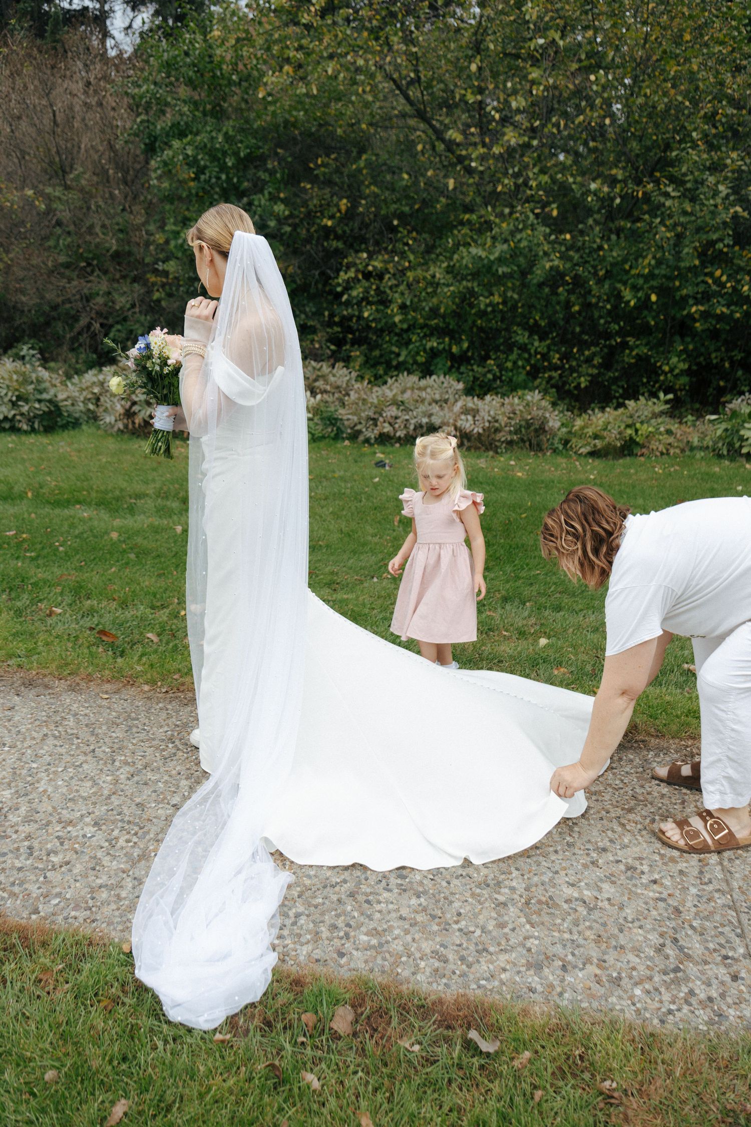 Flowing white wedding dress train trailing on gravel path with greenery in background.