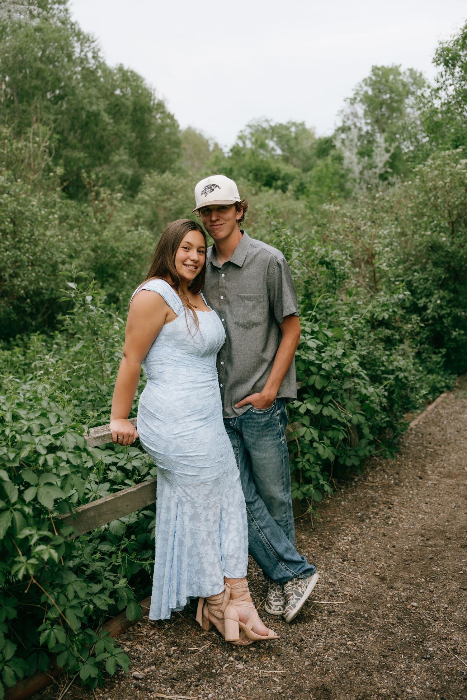 A couple poses together on a garden path surrounded by lush greenery, woman in blue lace dress and man in gray shirt.